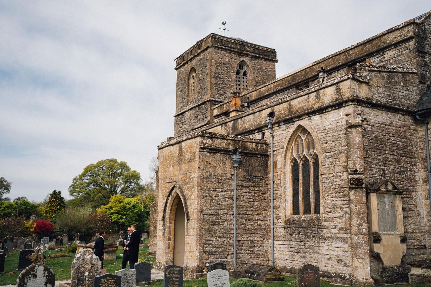 Historic stone church with gothic windows and tower, surrounded by an old cemetery with gravestones, under a cloudy sky.