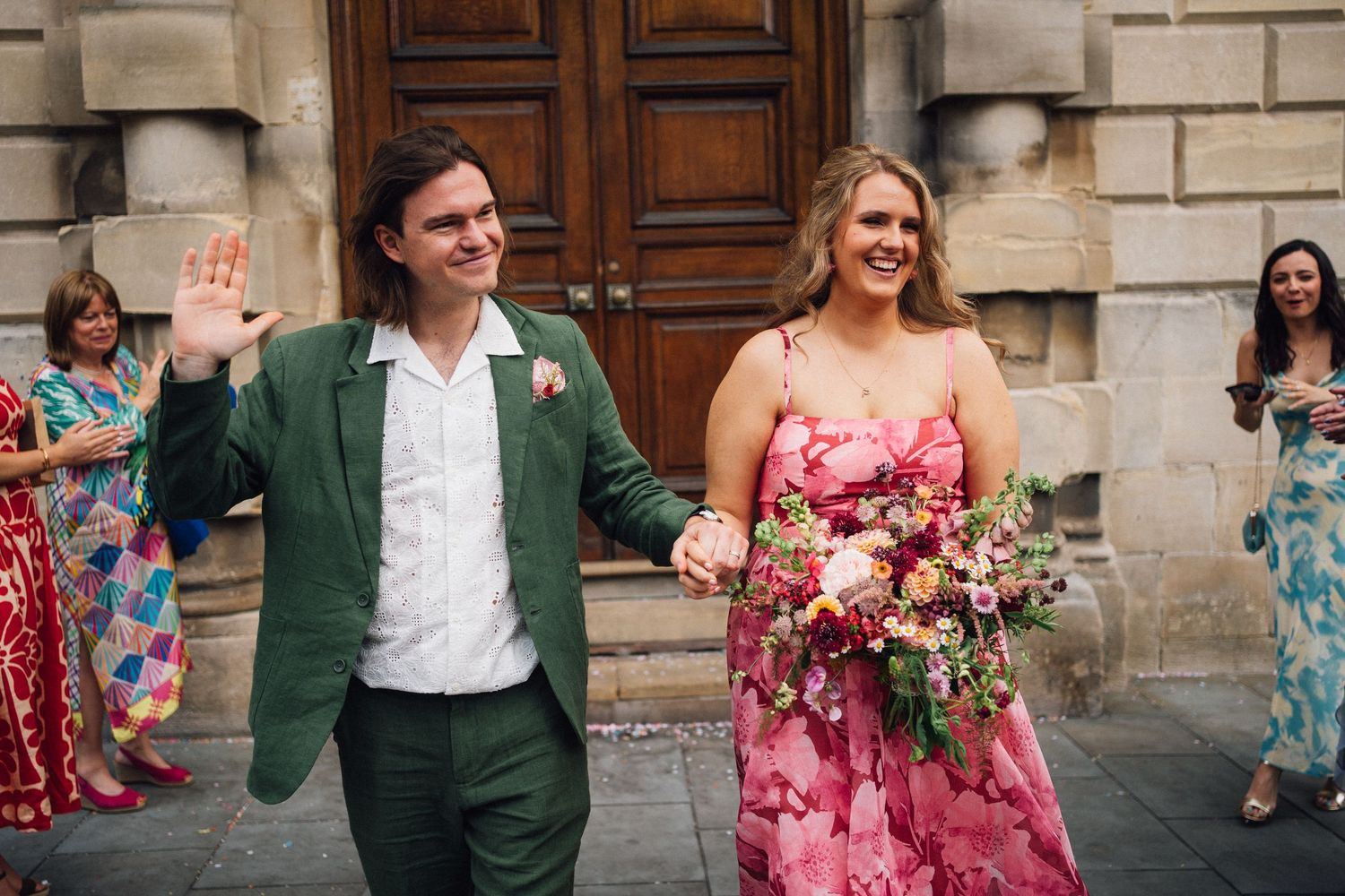 Newlywed couple walking out of ceremony venue, groom in green suit waving, bride in pink floral dress holding bouquet.