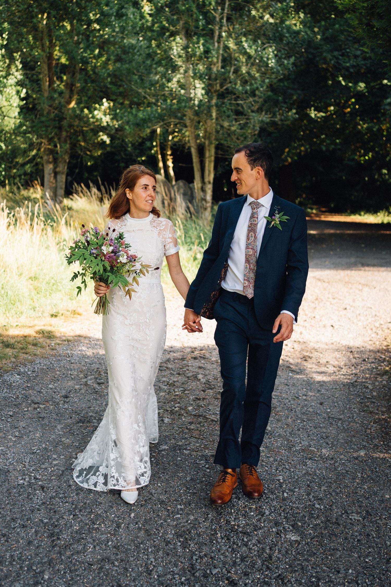 Bride and groom walk hand in hand on a gravel path, she in lace gown with bouquet, he in navy suit.