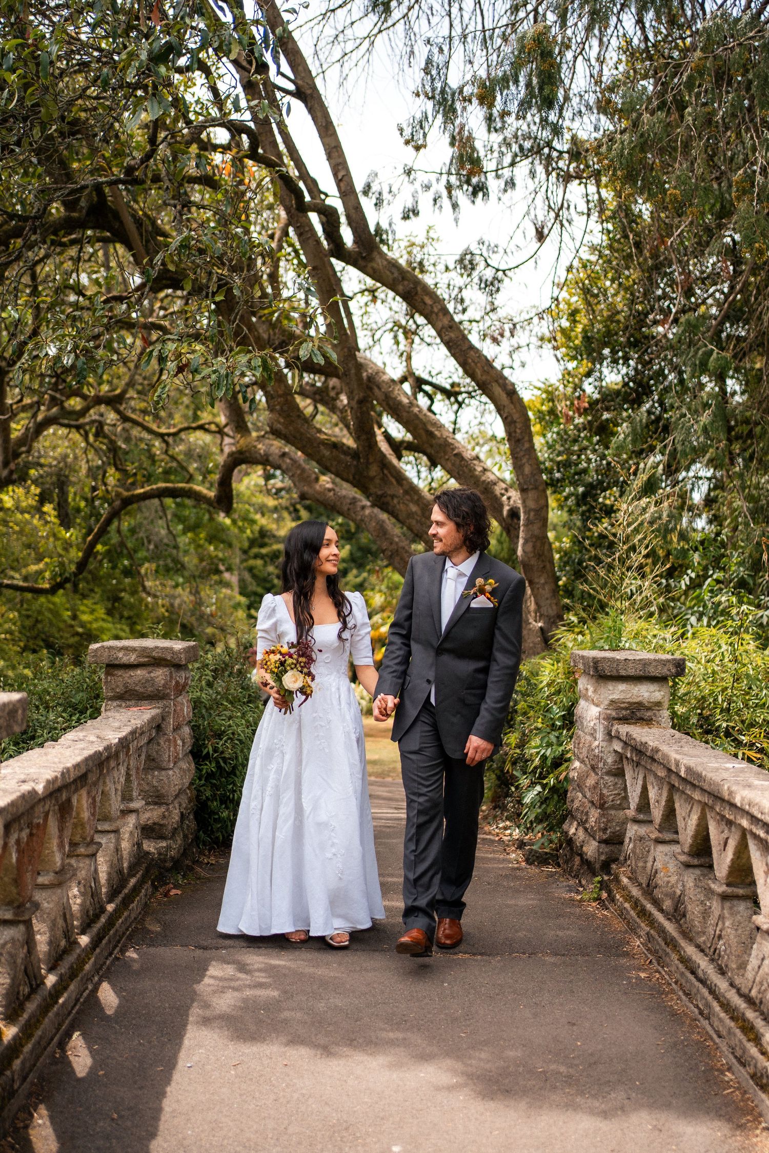 Bride and groom walking hand in hand on a stone bridge surrounded by lush green trees on their wedding day.