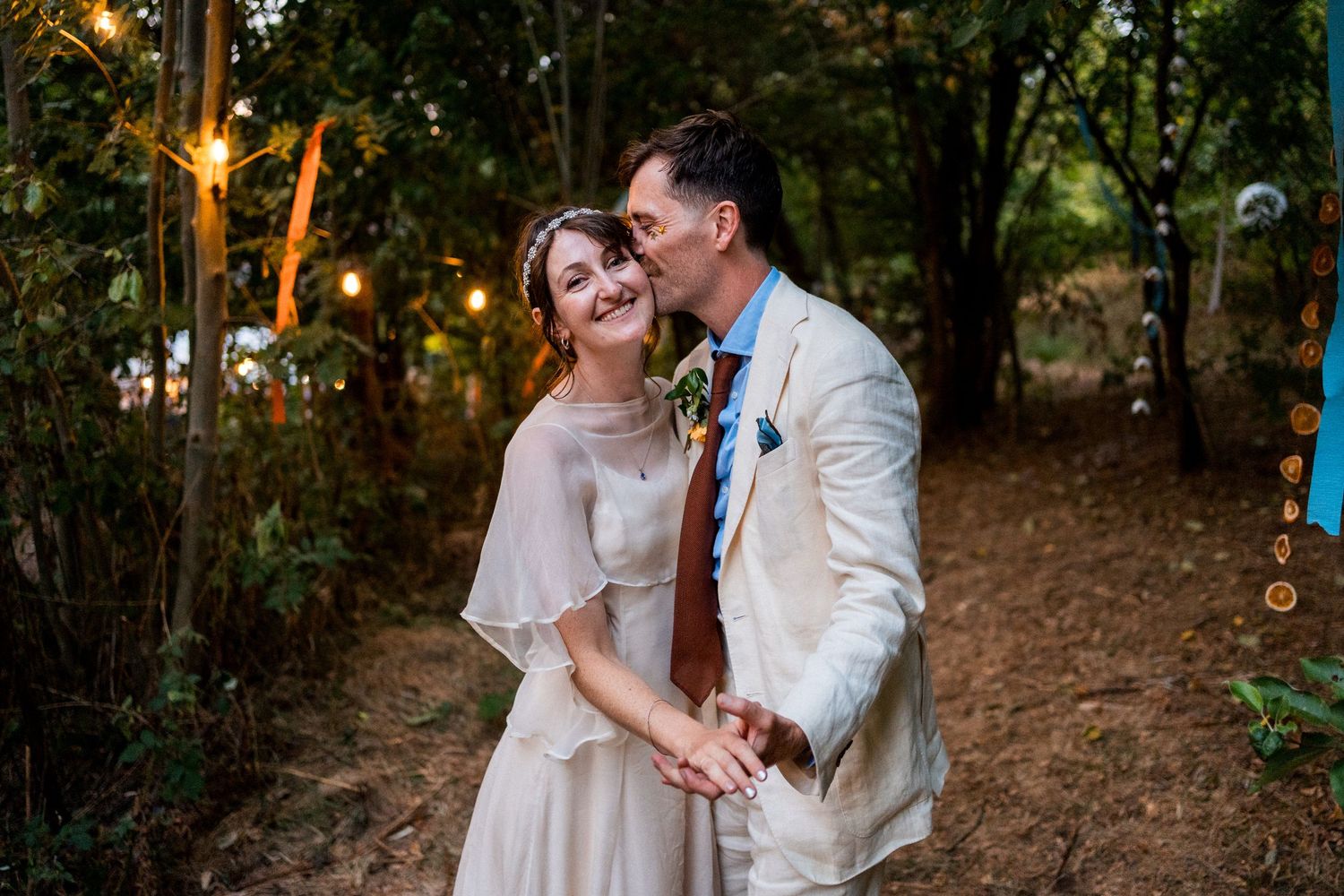 Couple shares a romantic kiss in a woodland wedding setting with string lights and lush greenery in the background.