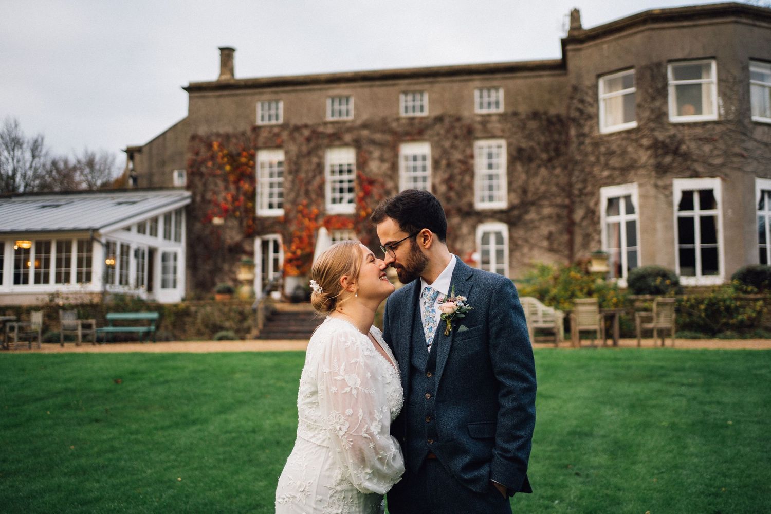 Bride and groom share a romantic moment on lush green lawn outside elegant stone manor house wedding venue.
