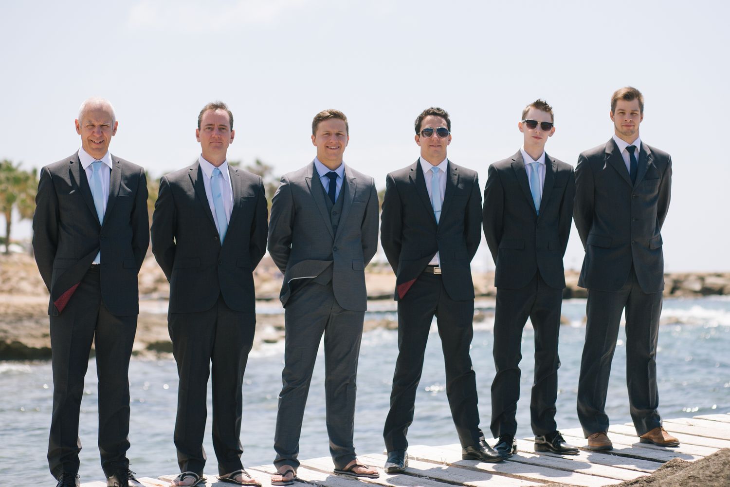 Groomsmen in black suits pose playfully on a beach boardwalk with water in the background.
