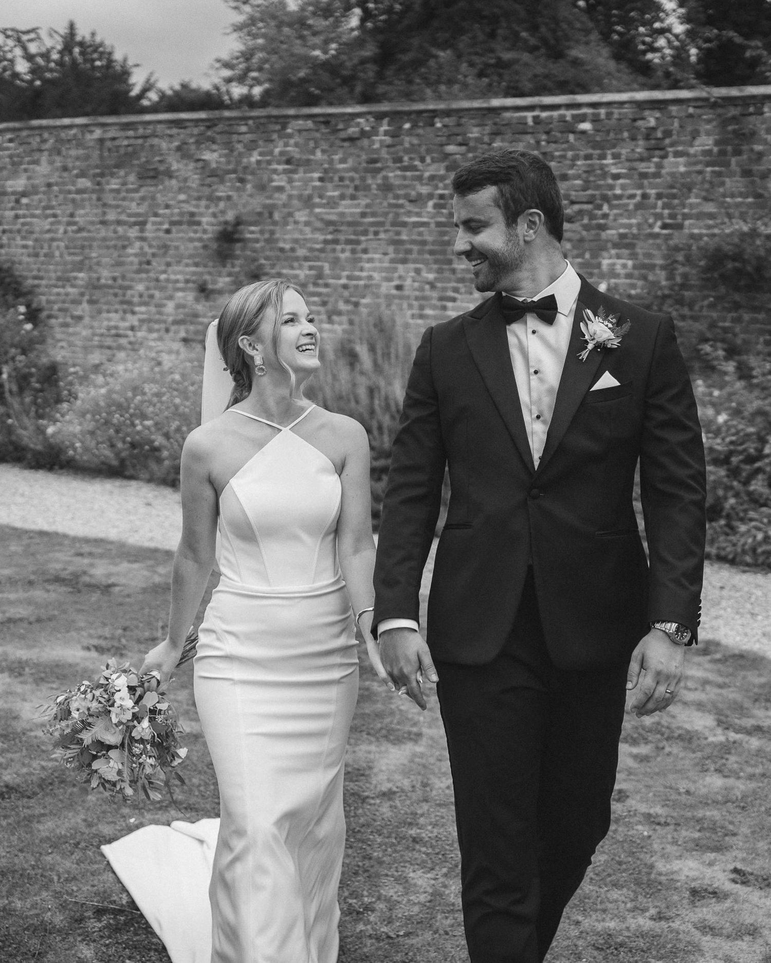 Wedding couple strolls hand in hand along a garden path with a classic brick wall background.