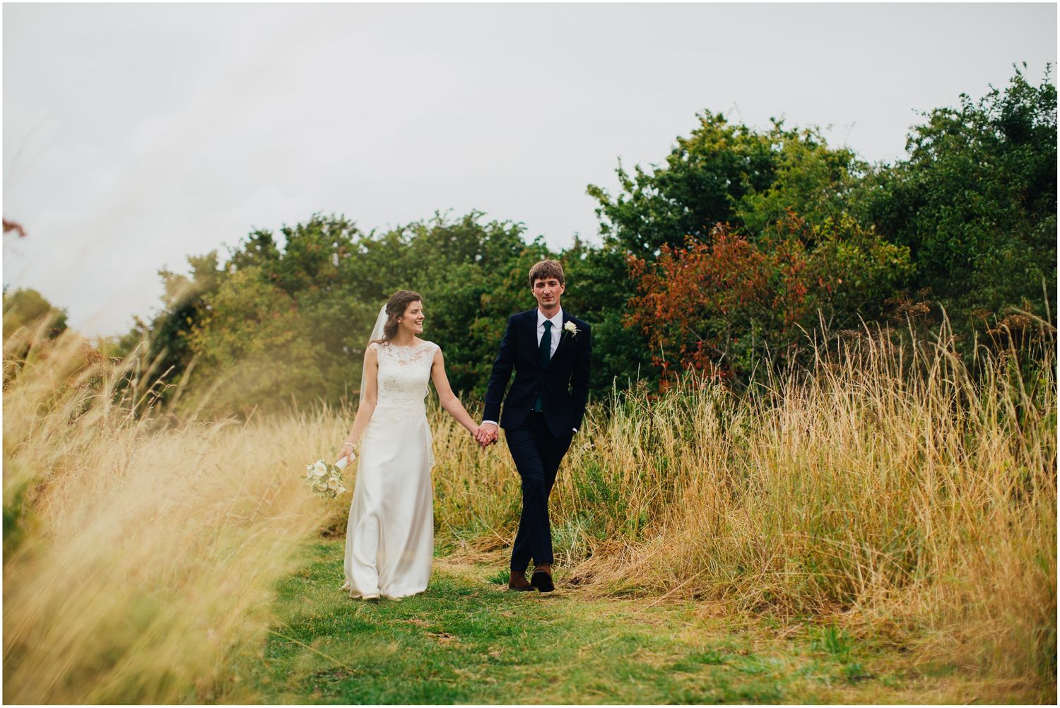 Wedding couple walks hand in hand through scenic meadow with tall grass.