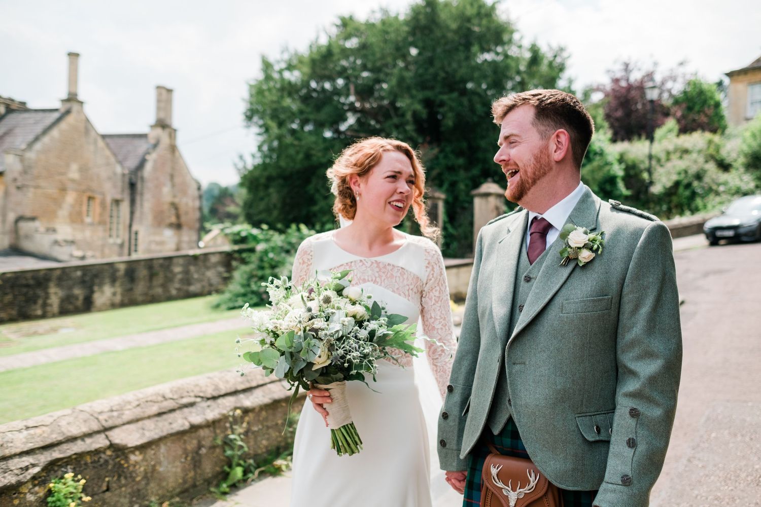 A couple laughing together outdoors on their wedding day near historic stone buildings.