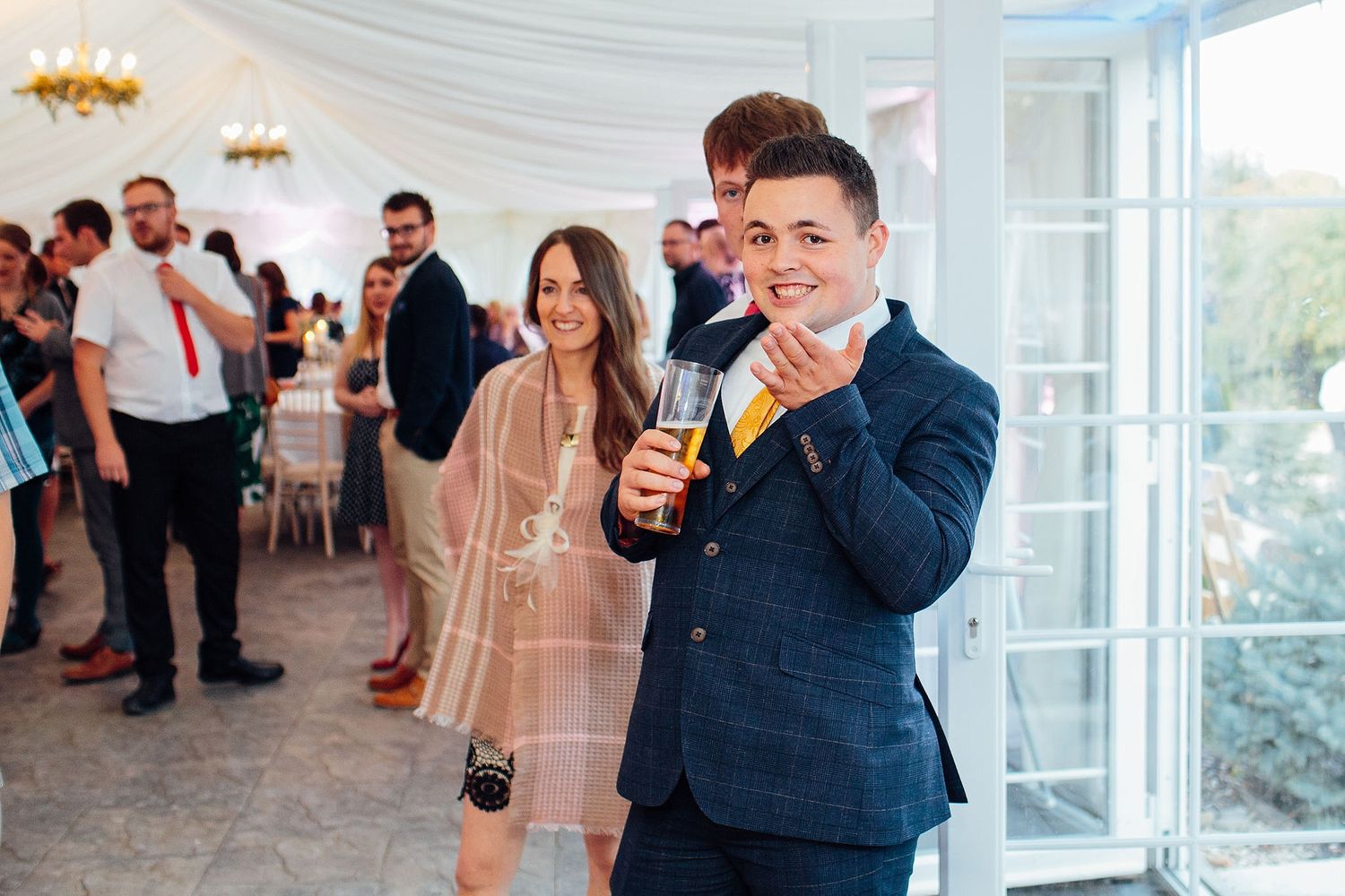 A guest in a navy suit enjoys a drink while celebrating at a wedding reception in a white tent venue.