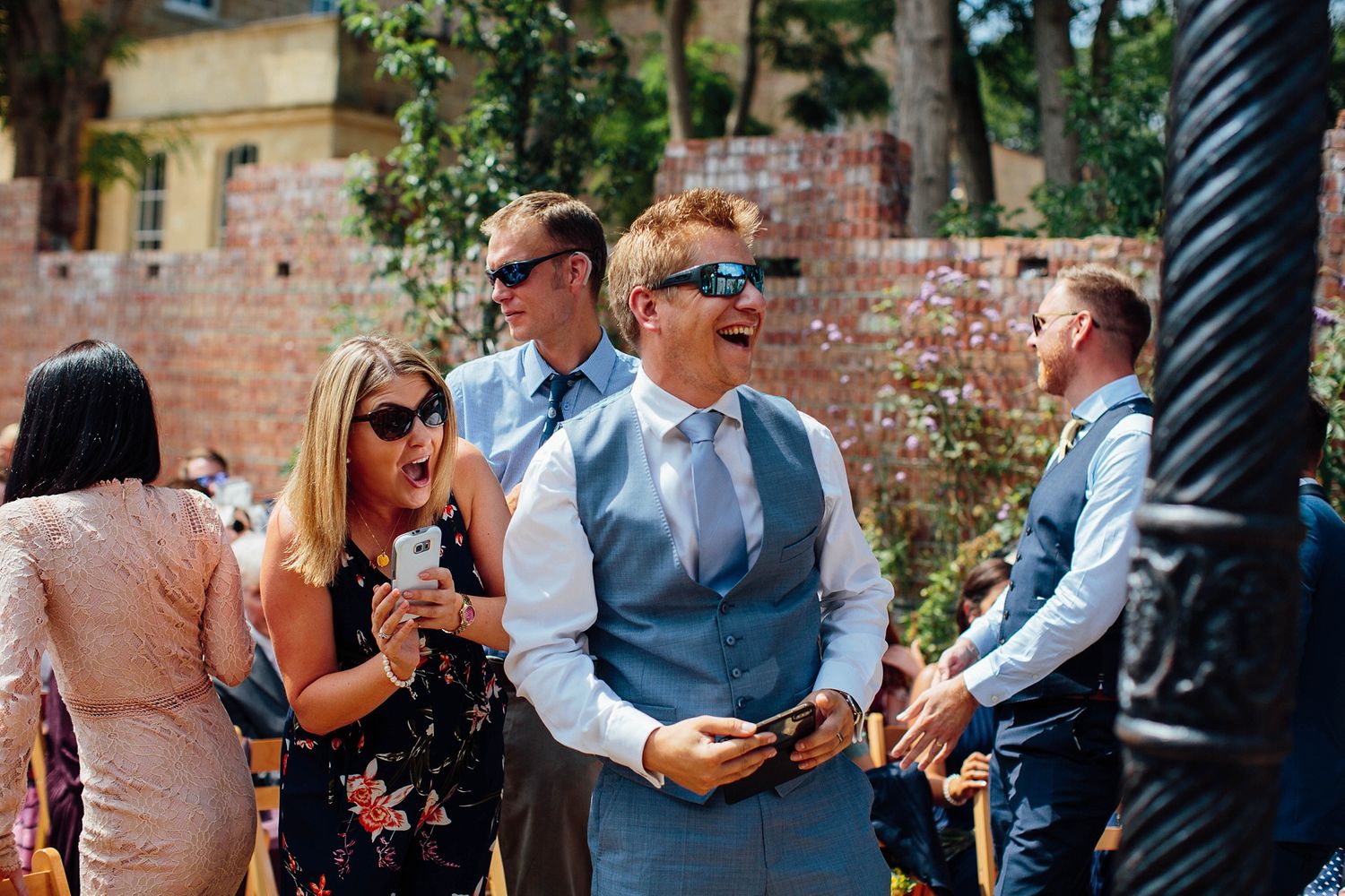 Wedding guests mingle during outdoor reception with brick walls and decorative lighting.
