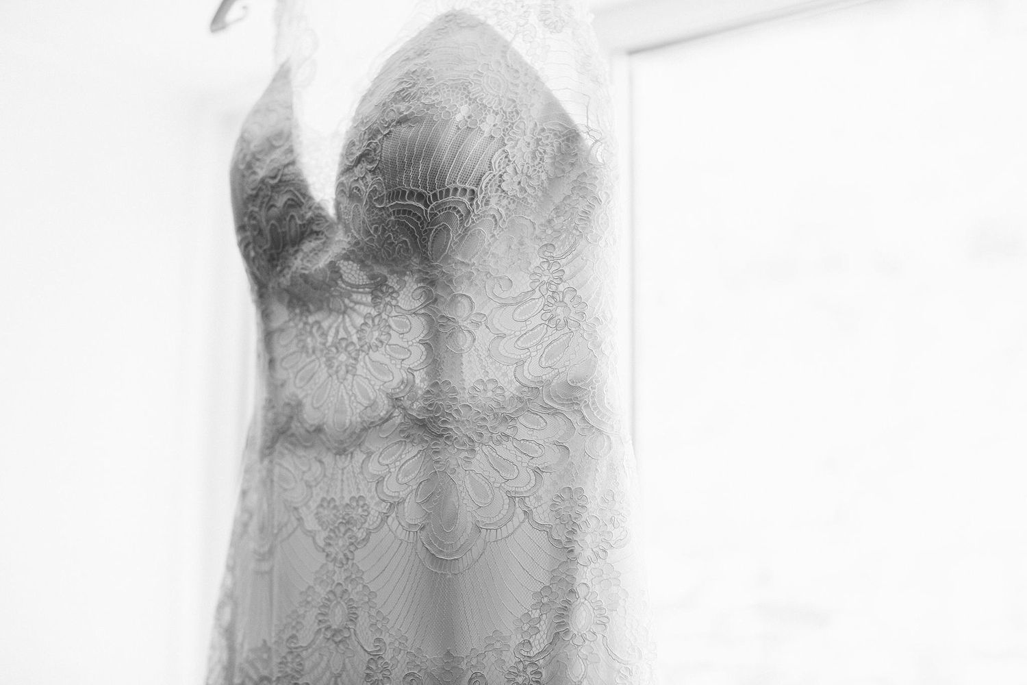 A delicate lace wedding dress hangs against a white background in black and white.