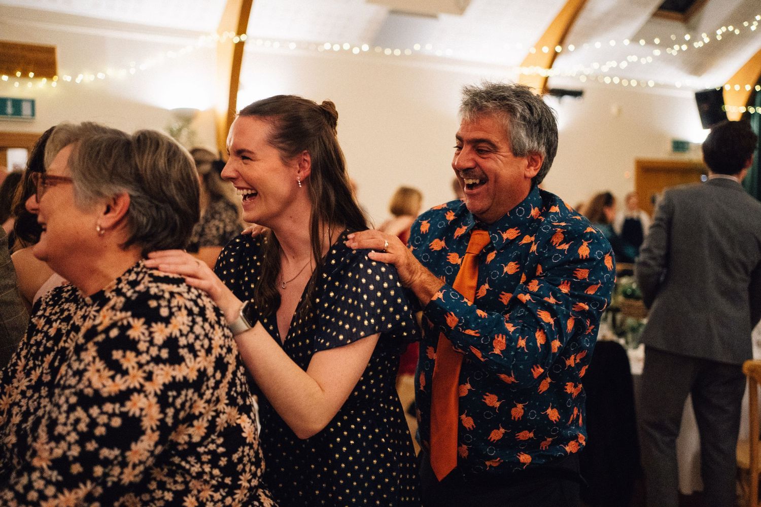 People laughing and socializing at an indoor event with string lights and wooden beams visible overhead.