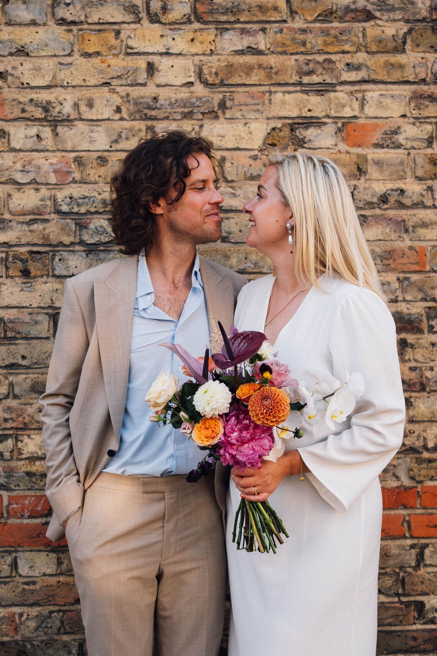 A couple in wedding attire share intimate moments against a rustic brick wall with a vibrant floral bouquet.