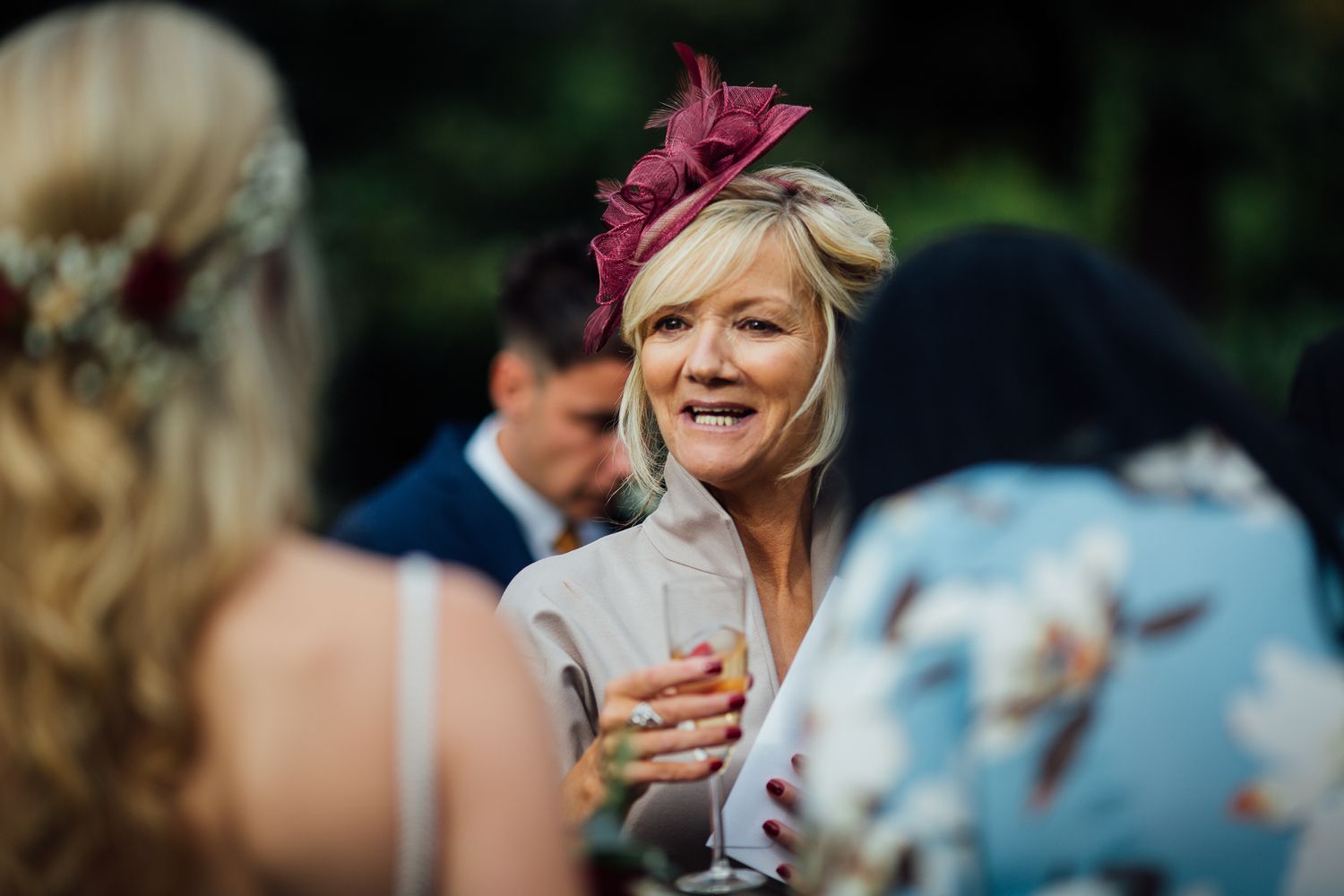 A guest in a burgundy fascinator enjoys a drink during an outdoor wedding reception.