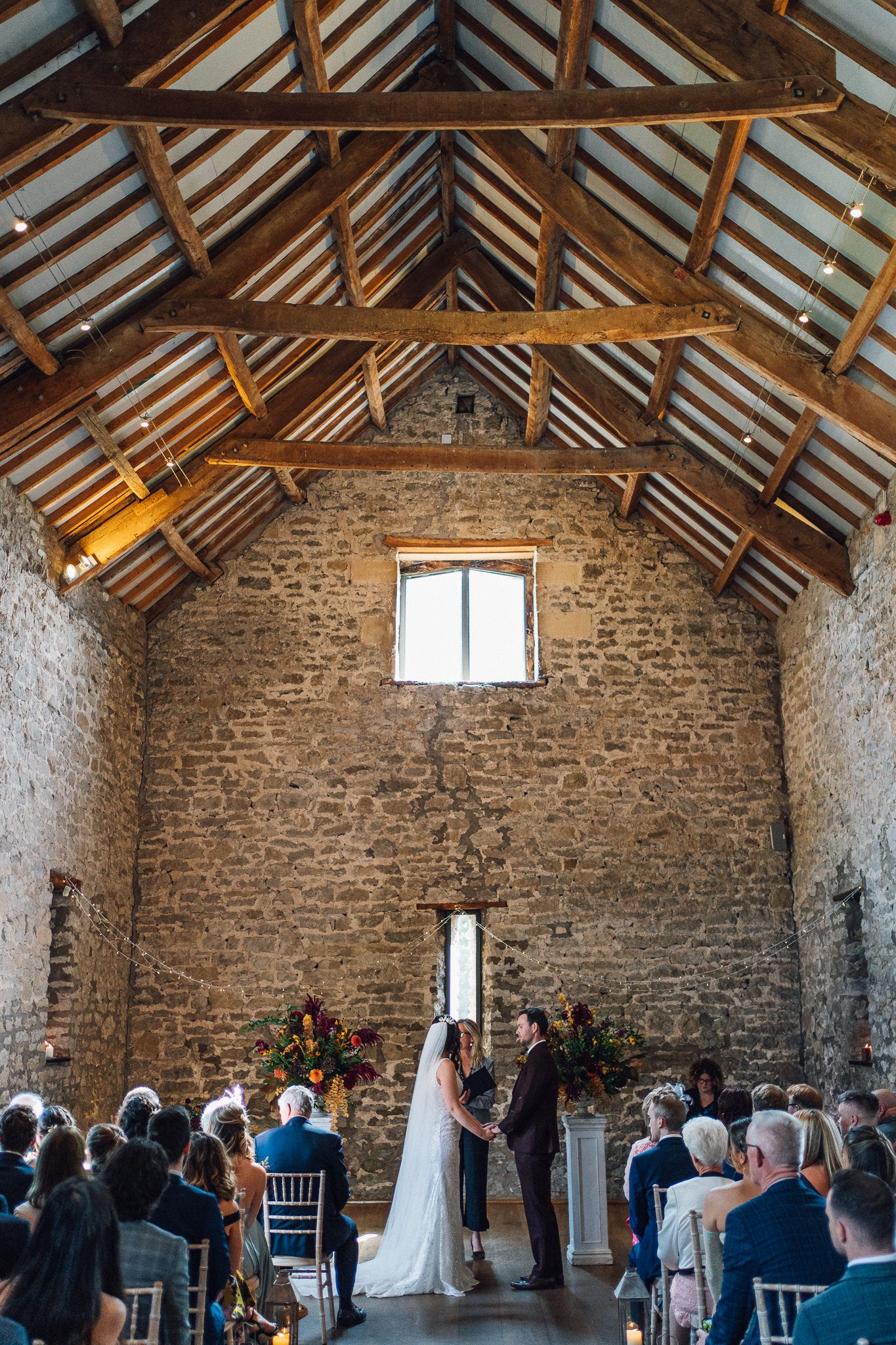 Indoor wedding ceremony taking place in rustic stone barn with exposed wooden beams and natural lighting.