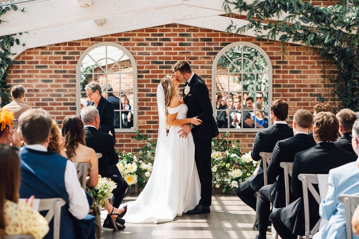Intimate outdoor wedding ceremony moment under an elegant brick pergola surrounded by seated guests.