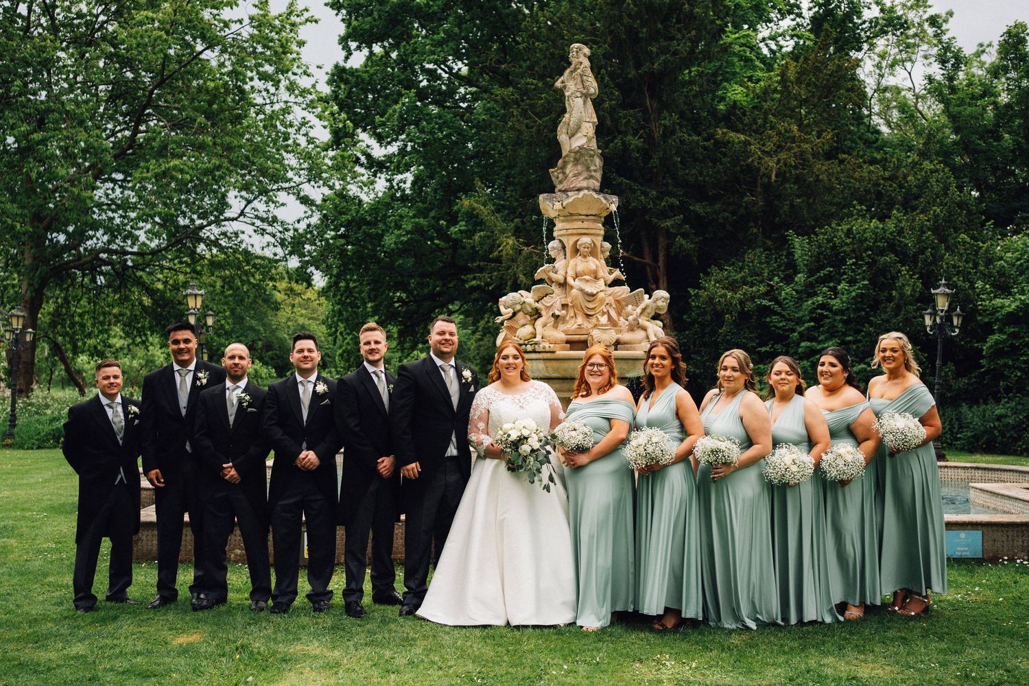 Wedding party in sage green dresses and black suits pose together on a grassy lawn near stone pillars.