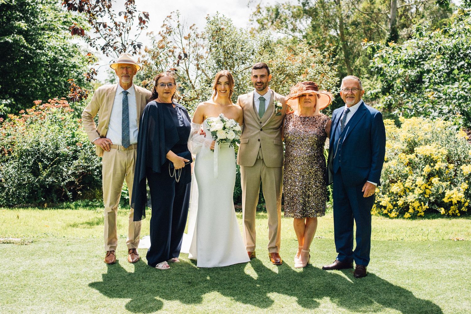 Wedding party gathered outdoors on a sunny day in a garden setting with yellow flowers in the background.
