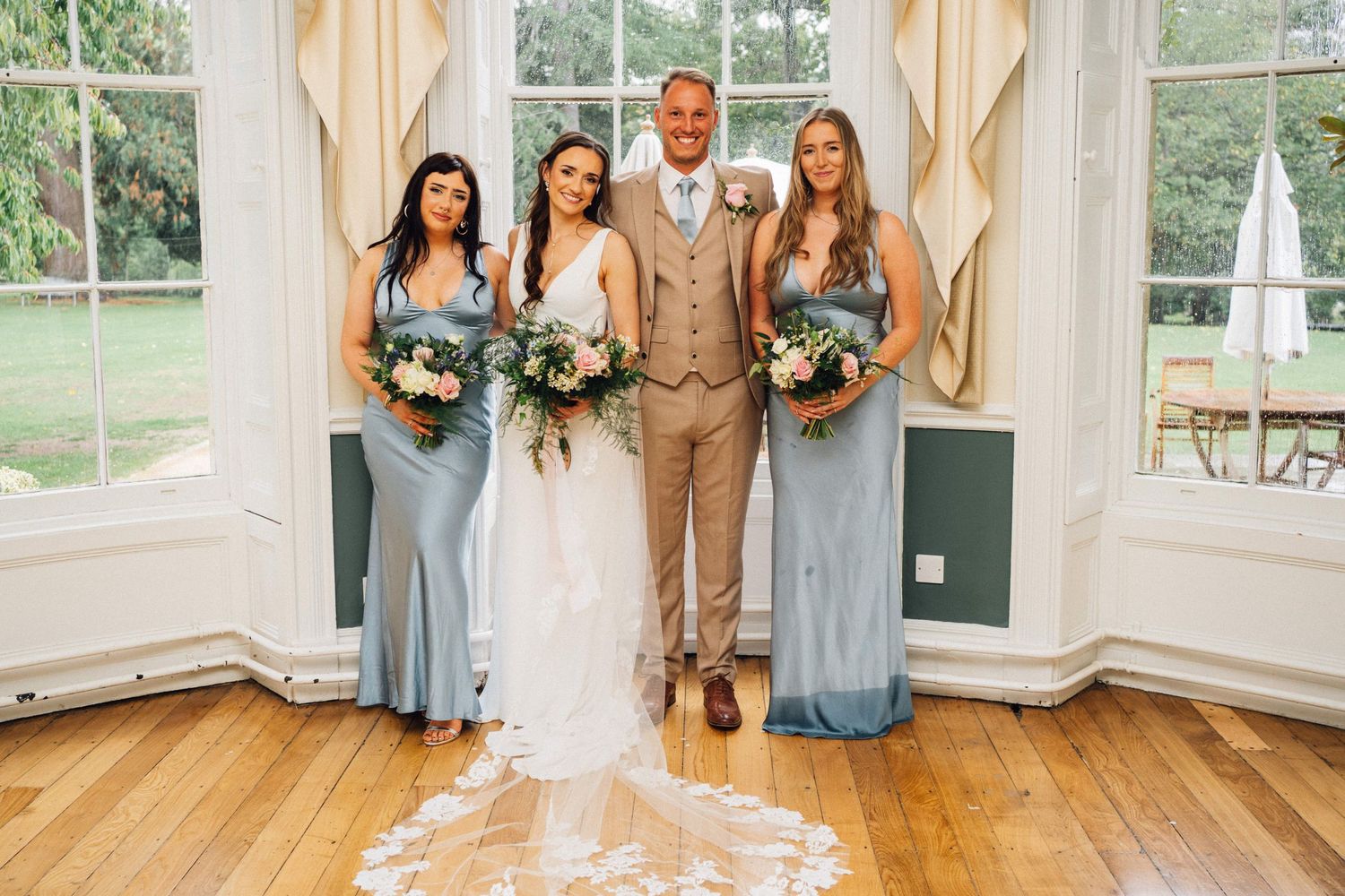 Wedding party members arranged for formal group photos in a sunlit room with hardwood floors.