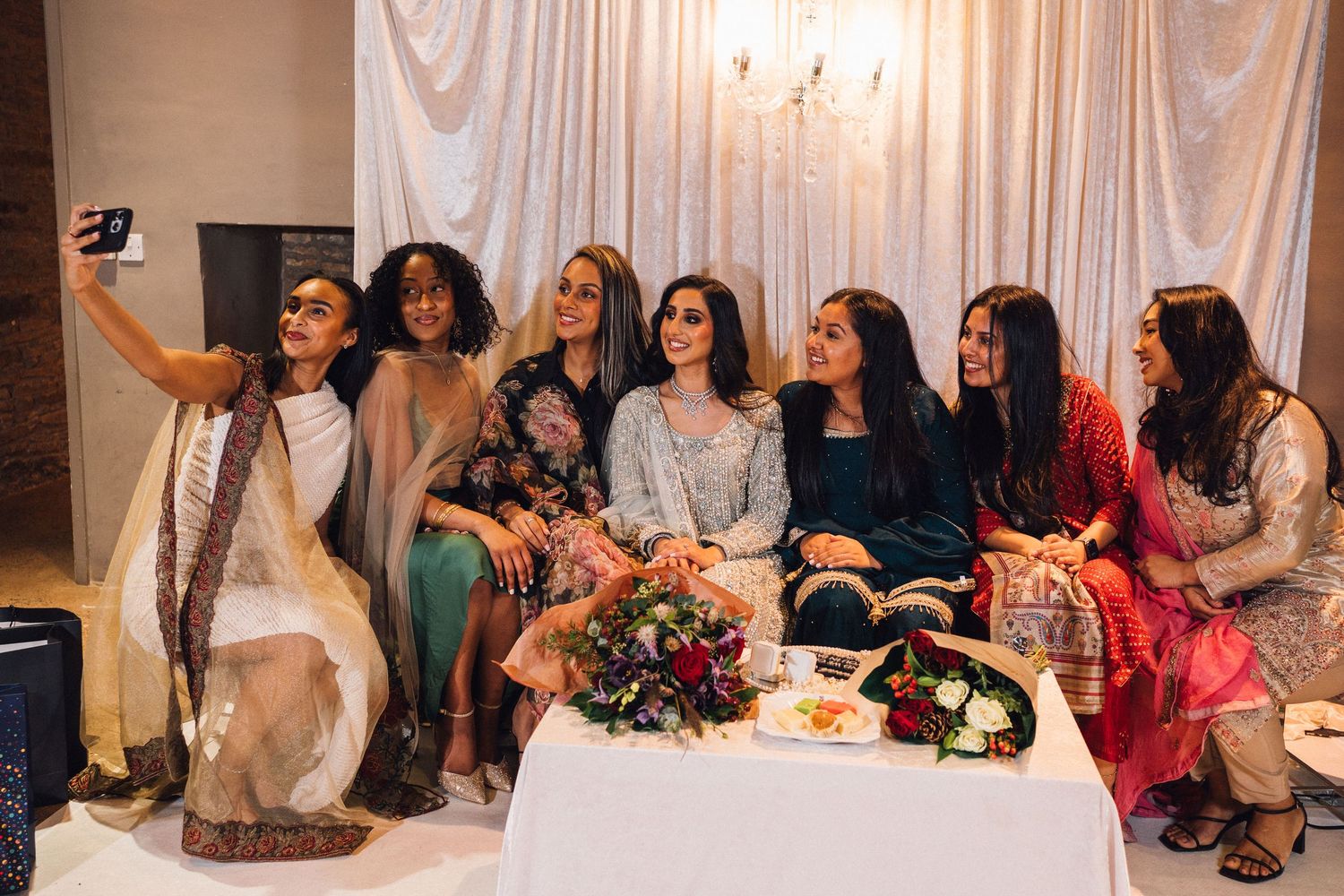 Wedding celebration guests gathered around dining tables with decorative floral arrangements and white backdrop.