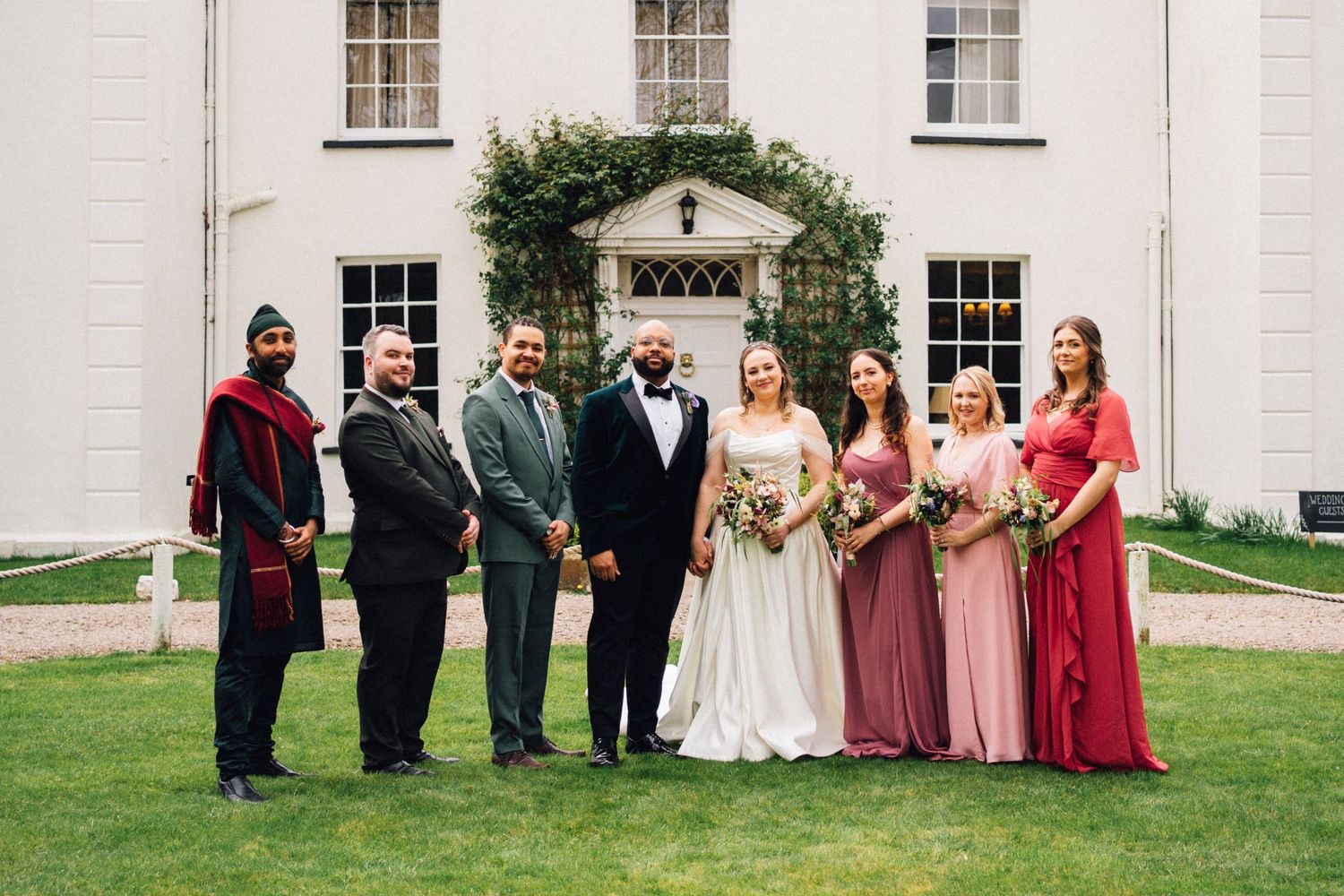 Wedding party poses in front of a white church building with pink and burgundy bridesmaids dresses.