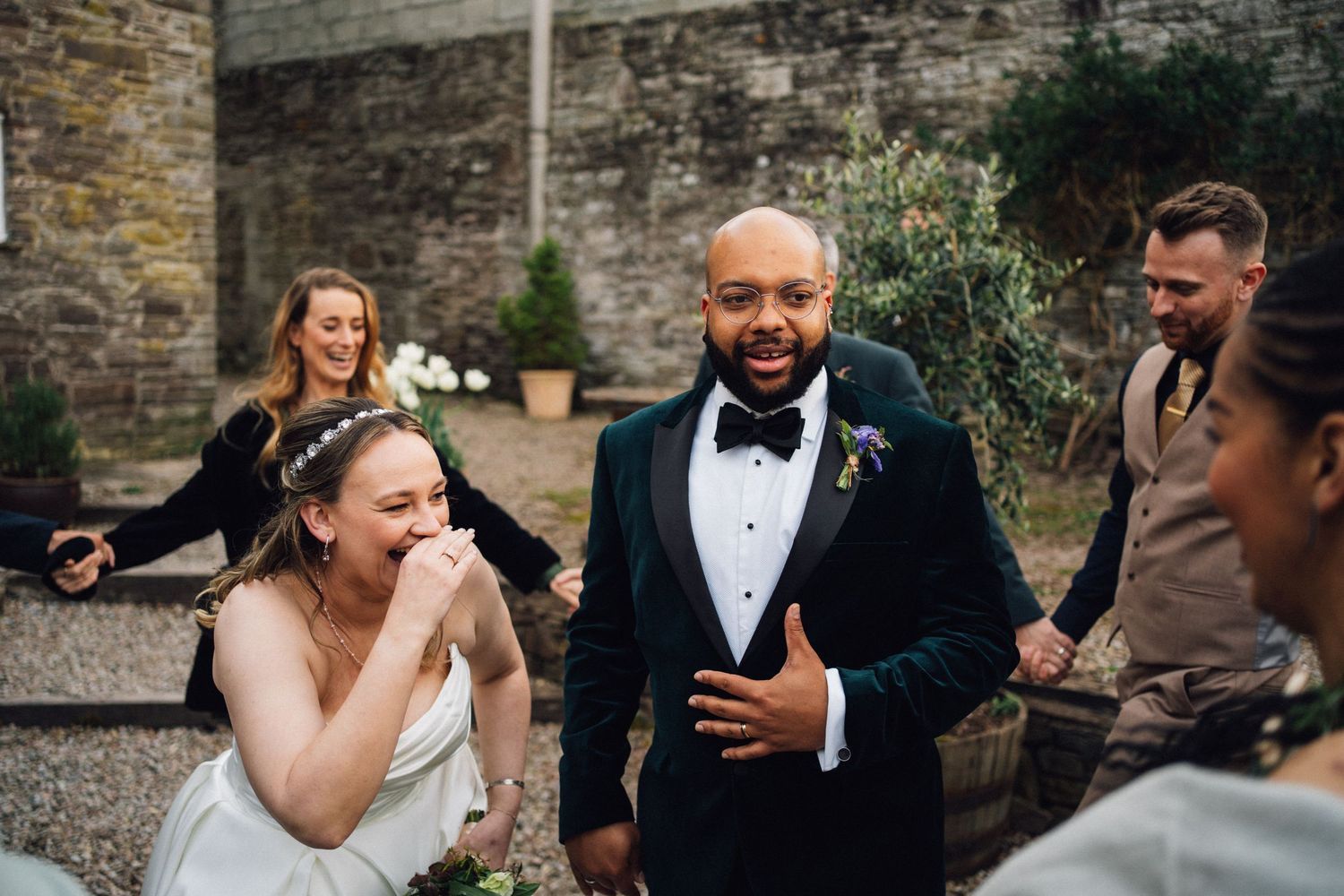 Wedding celebration candid moment outside stone walls with guests laughing together.