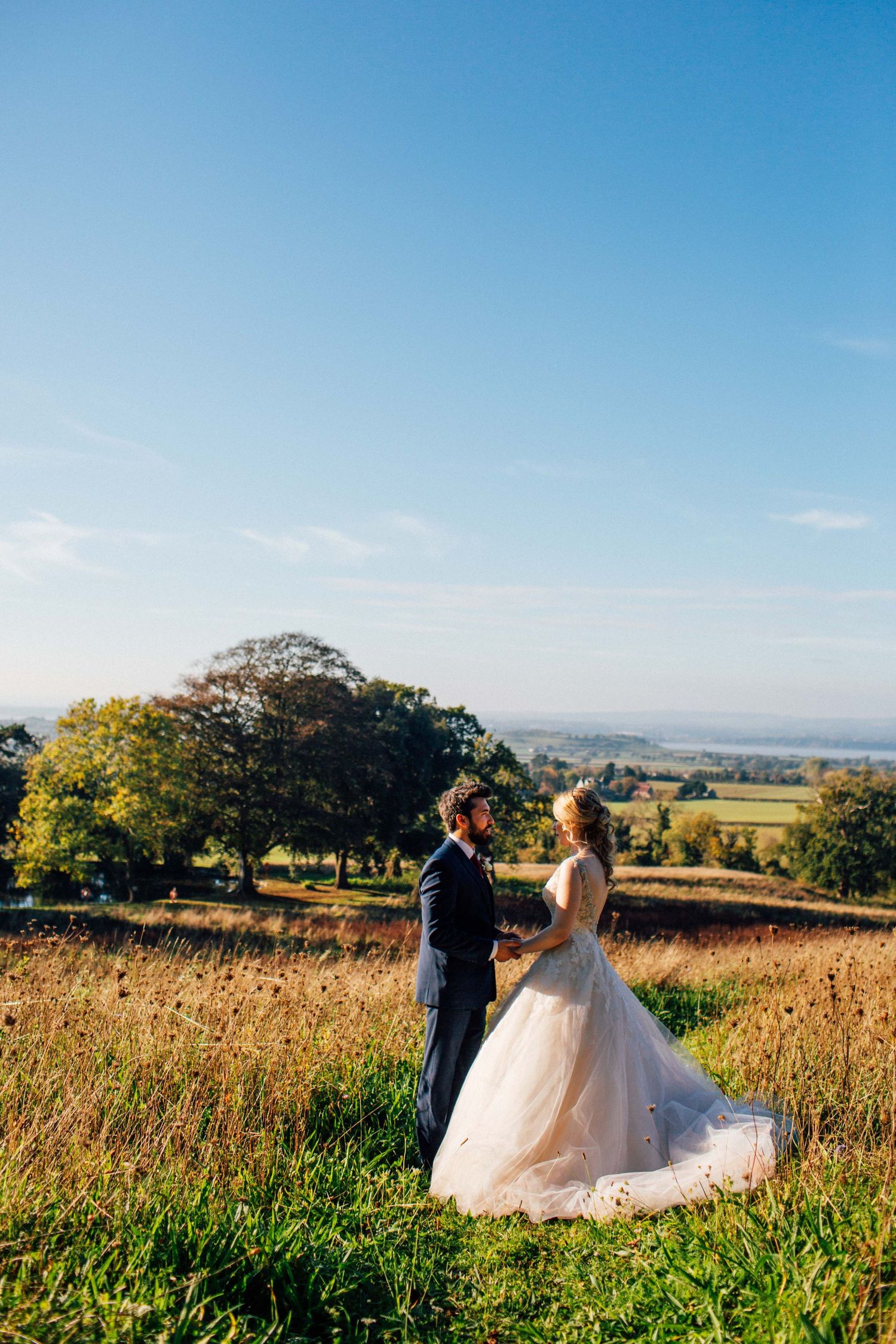 A couple in wedding attire walks through a scenic golden meadow under bright blue skies on their special day.