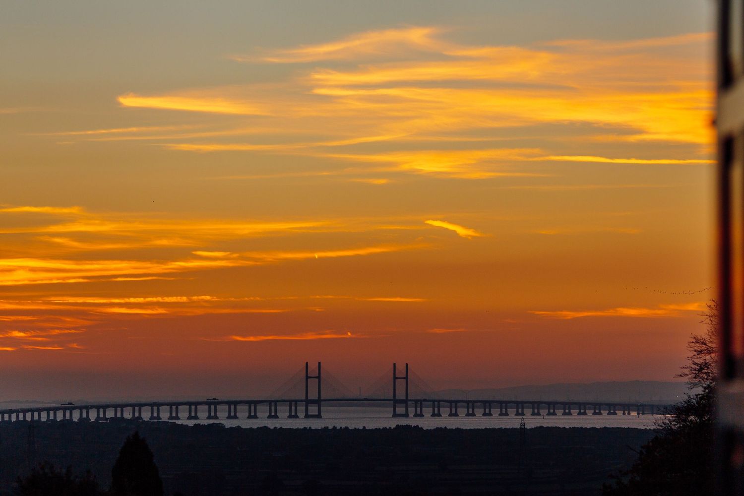 Dramatic orange sunset sky silhouettes a bridge and coastline against vibrant dusk colors.