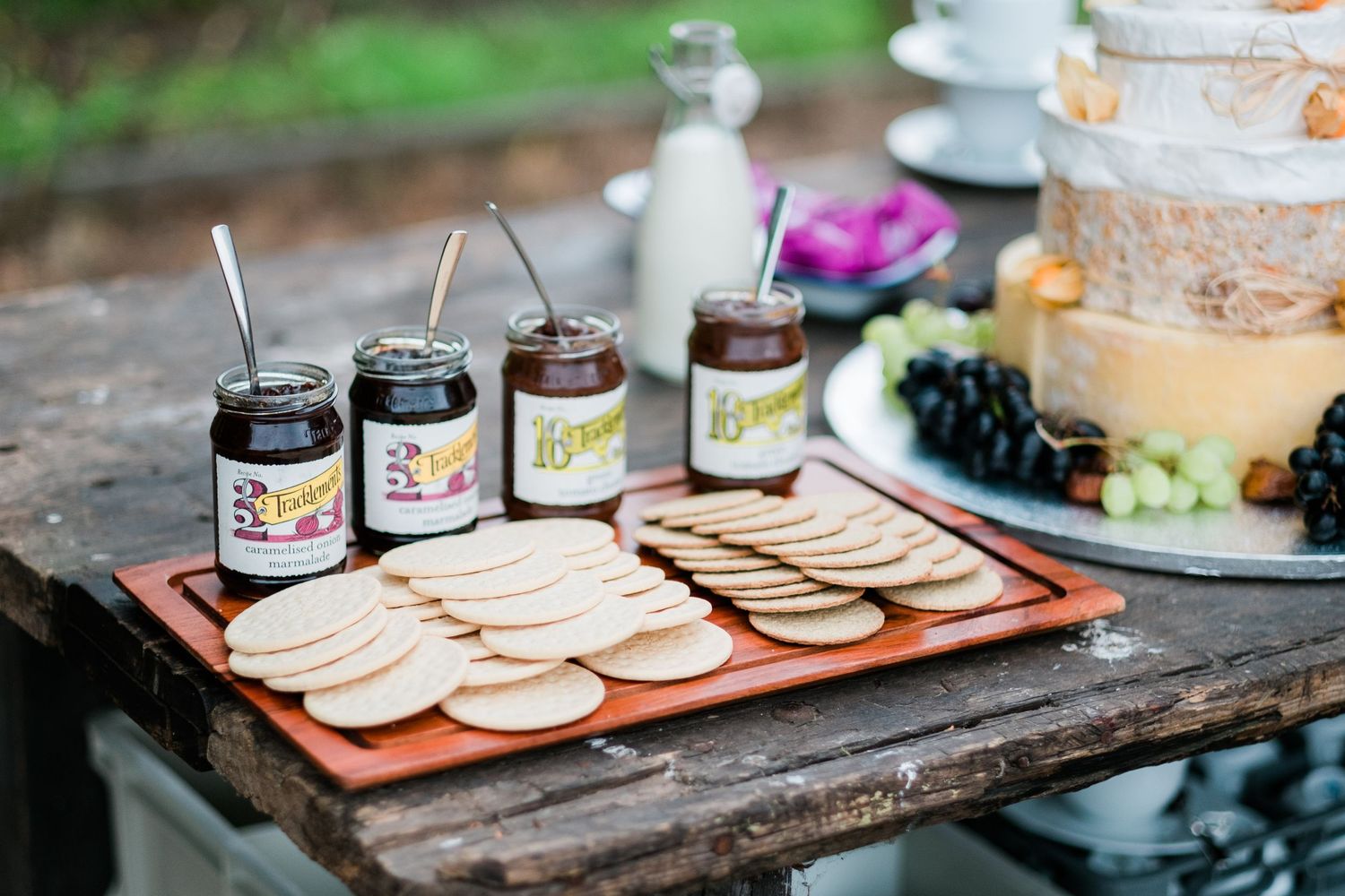 Wooden serving board with crackers and jars of spreads arranged for an outdoor gathering.