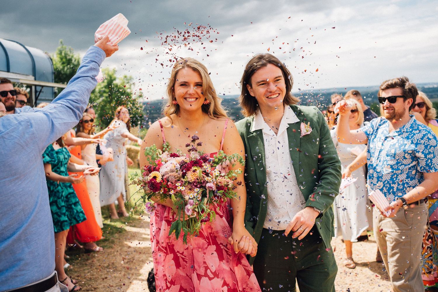 Wedding guests celebrate with confetti and flowers during an outdoor celebration under cloudy skies in Kelston Roundhill the Old Barn
