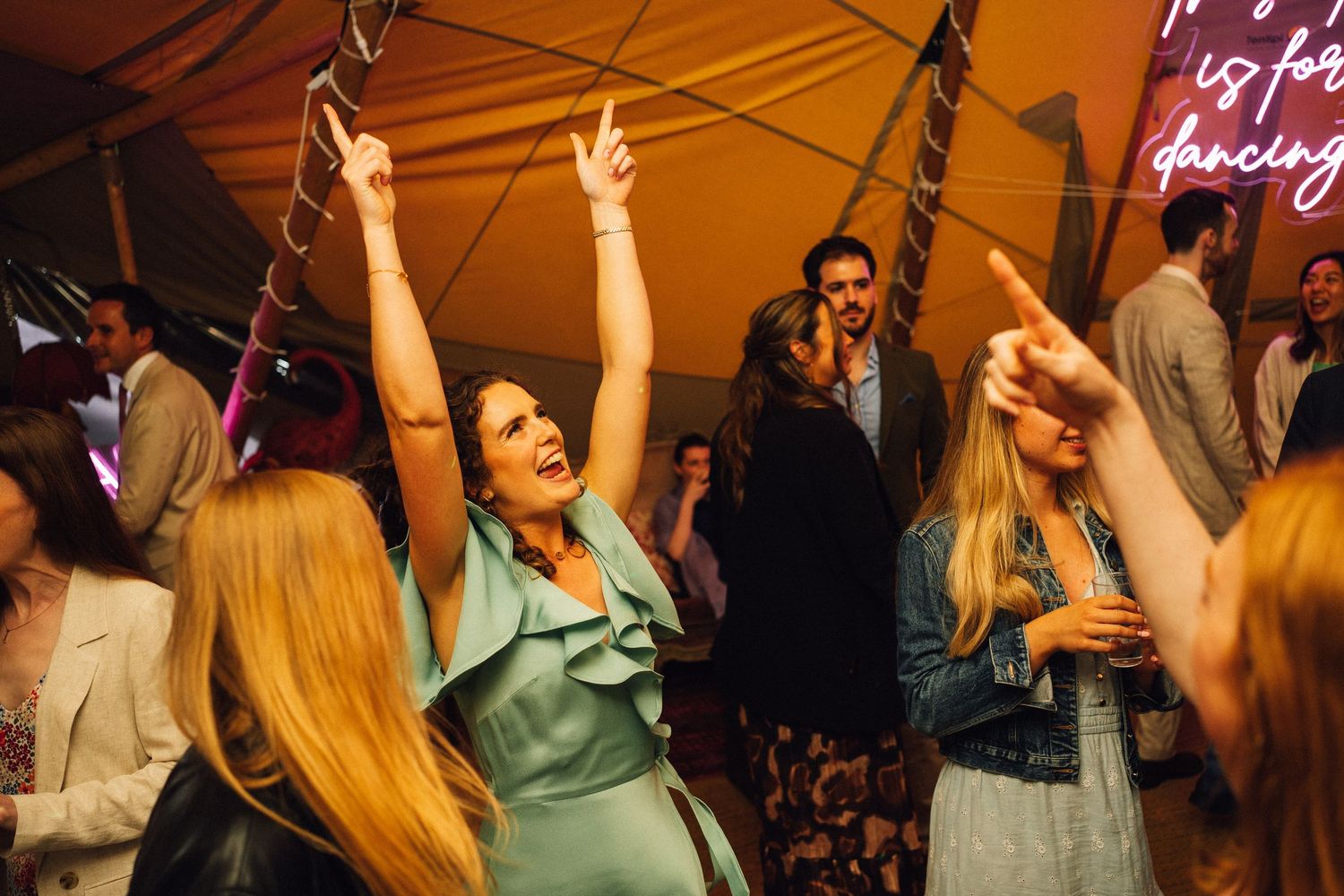 People dancing enthusiastically with raised arms on a dance floor illuminated by warm lighting and neon signs.