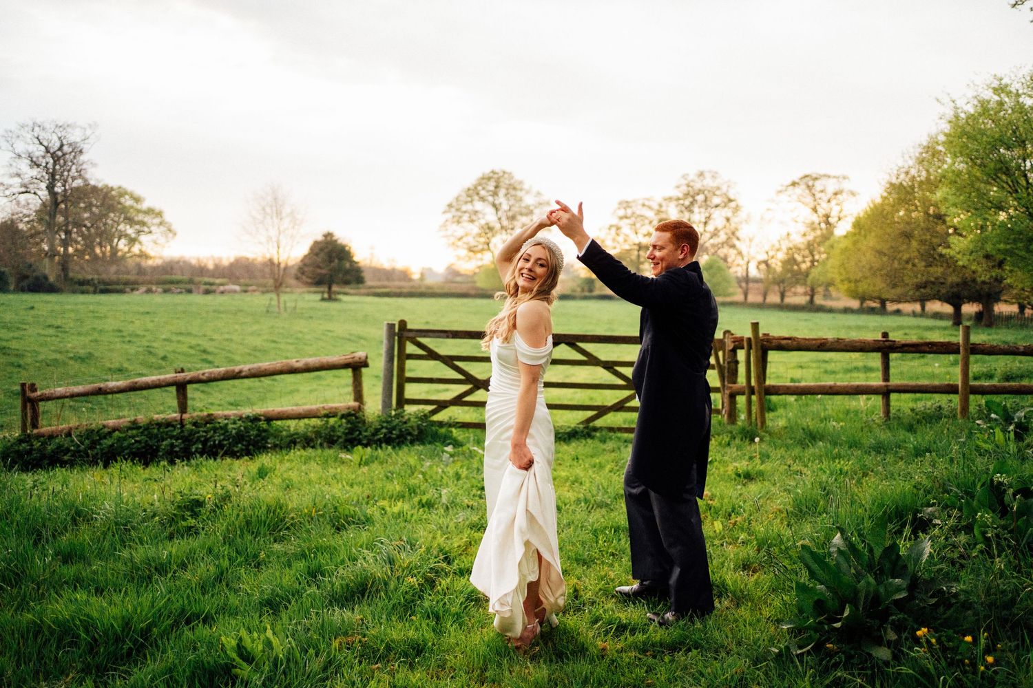 A couple walks hand in hand through a scenic countryside setting with wooden fences and green fields at sunset.