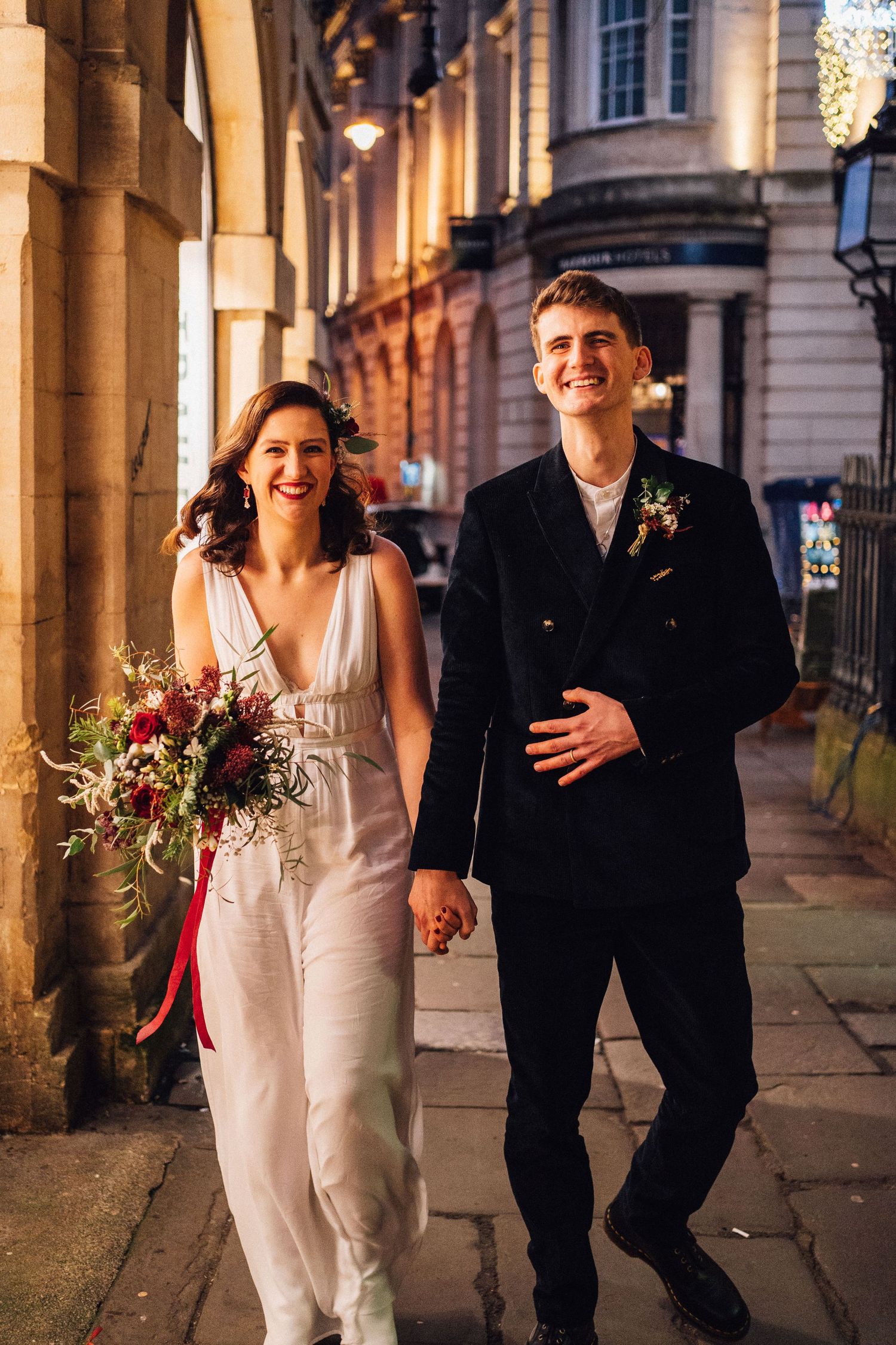 A bride and groom share joyful moments together while walking under illuminated archways at night.