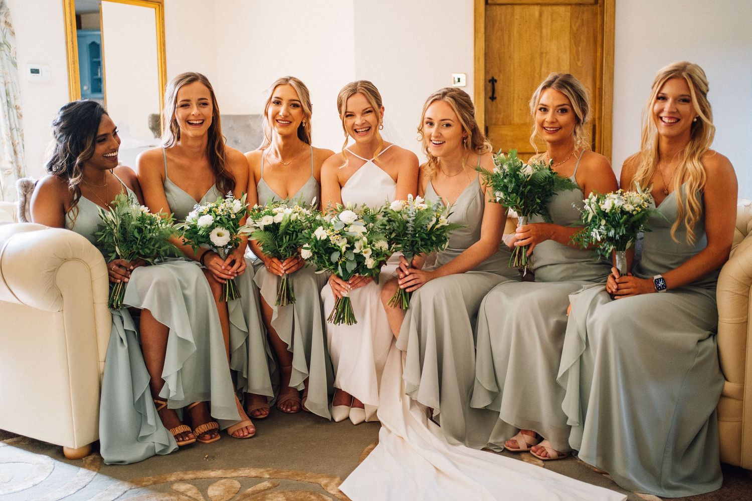 Bridesmaids in sage green dresses pose together with white floral bouquets at an elegant wedding celebration.