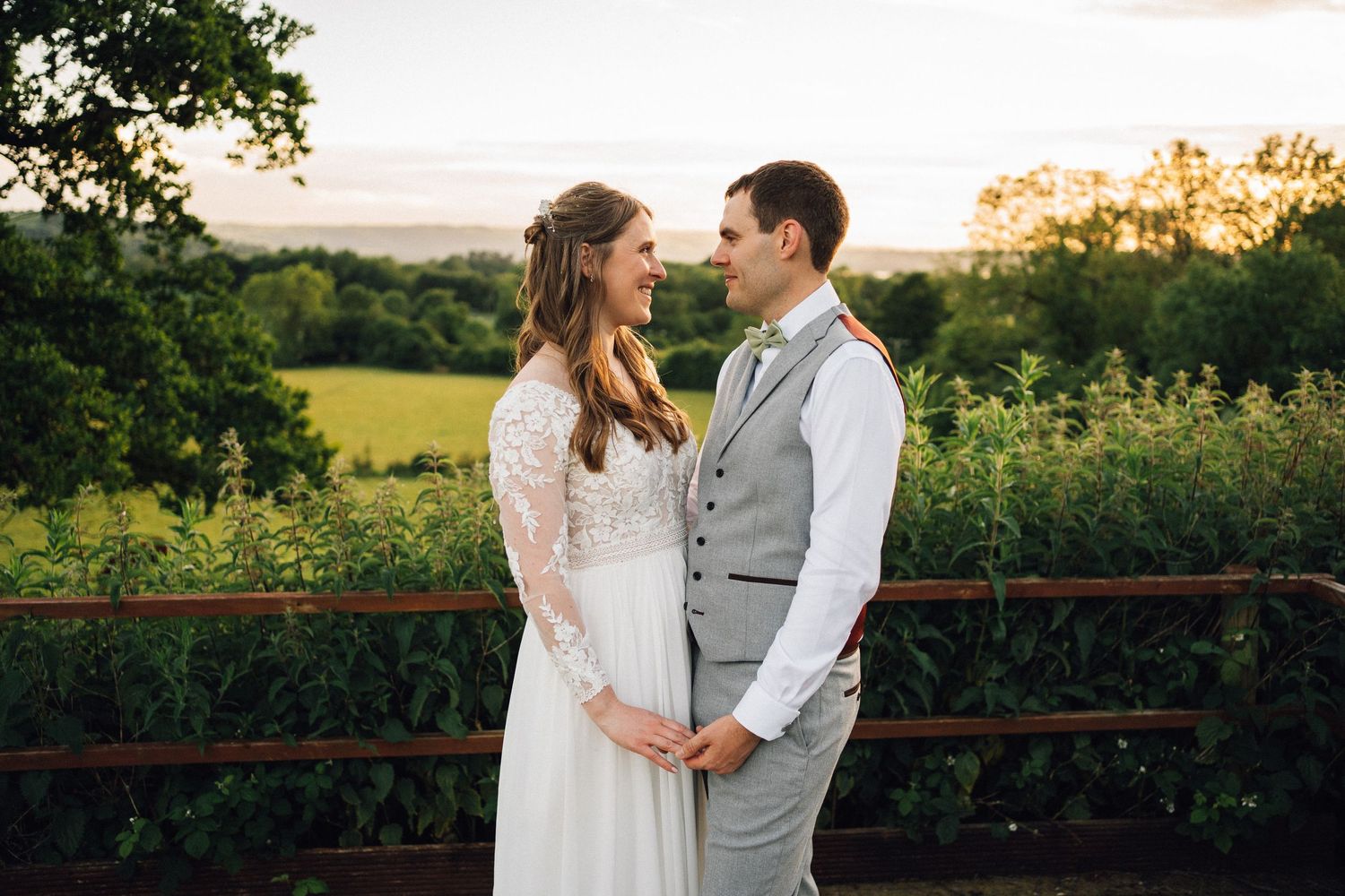 Bride and groom share a romantic moment at sunset overlooking lush green countryside fields on their wedding day.