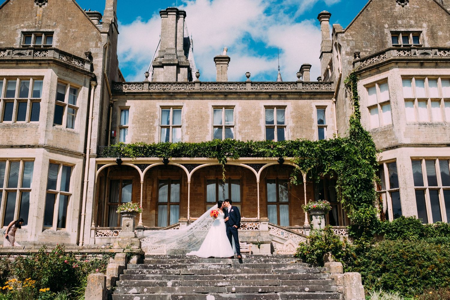 Bride and groom stand on grand stone steps of an elegant English manor estate on a sunny wedding day.