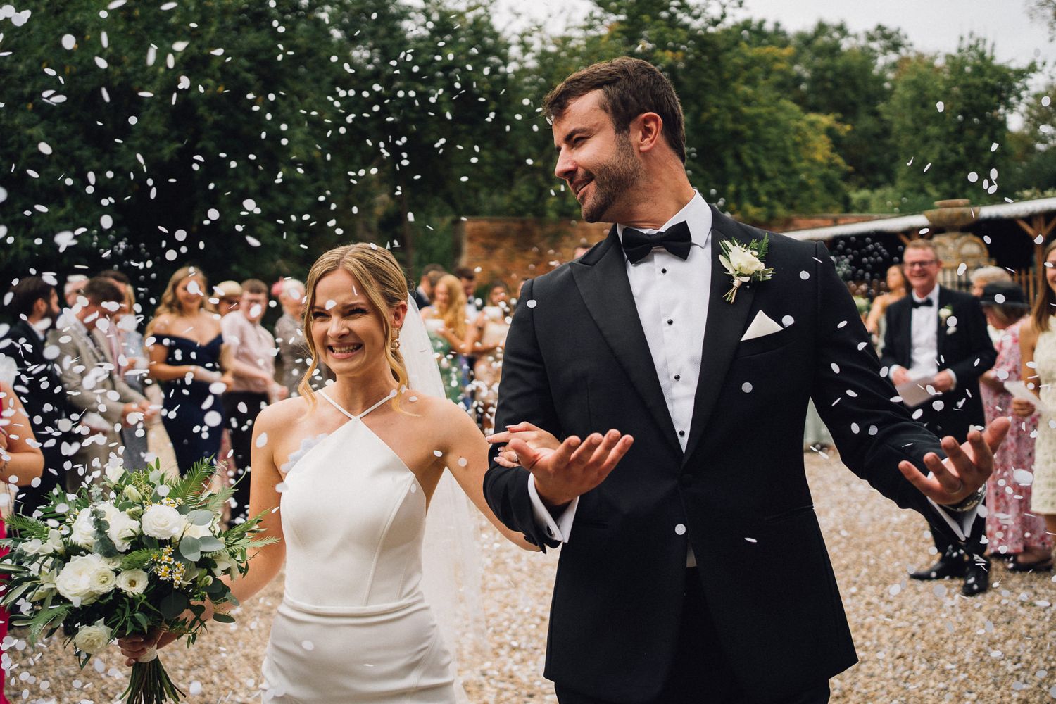 Wedding couple walking through confetti shower with guests celebrating outdoors.