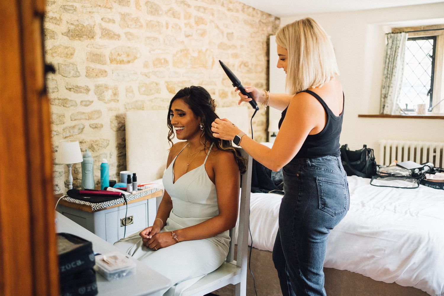 A stylist works on hair preparation in a rustic stone-walled room with natural lighting.