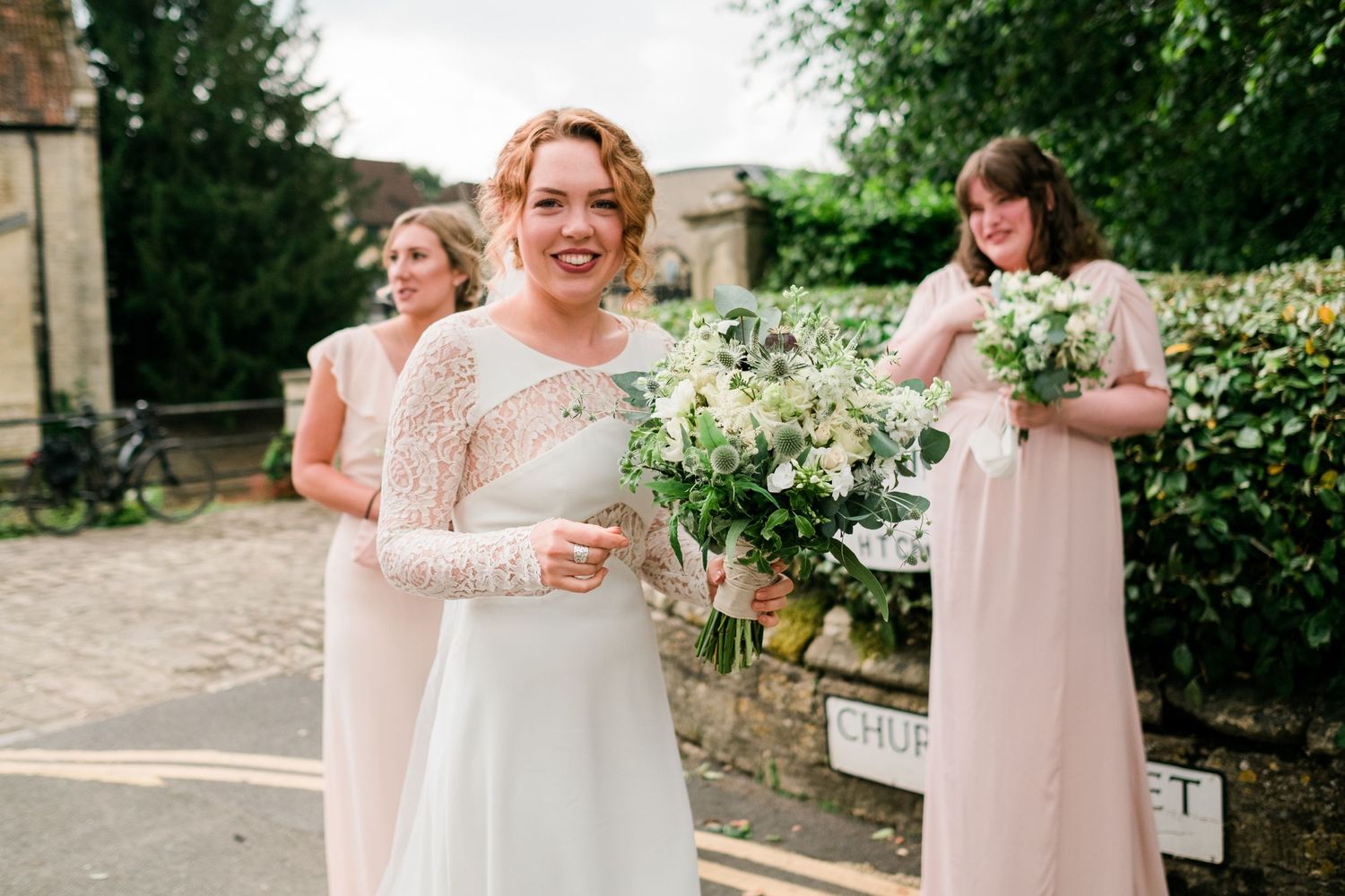 Wedding party members in light colored dresses gathered outdoors near flowering bushes.