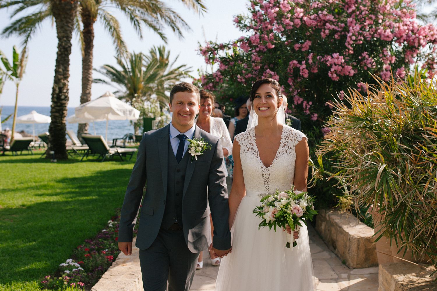 Newlyweds walking hand in hand through a tropical garden with palm trees and pink flowers.