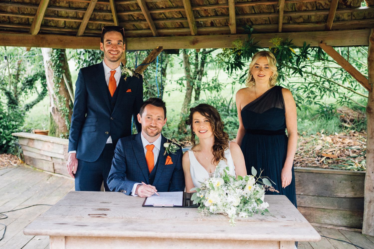 Wedding couple signs documents under a rustic wooden pavilion during ceremony.