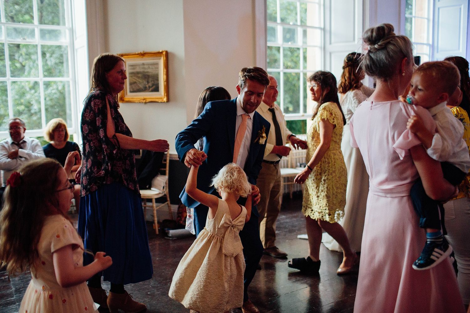 Wedding reception guests dancing and socializing in a bright room with large windows.