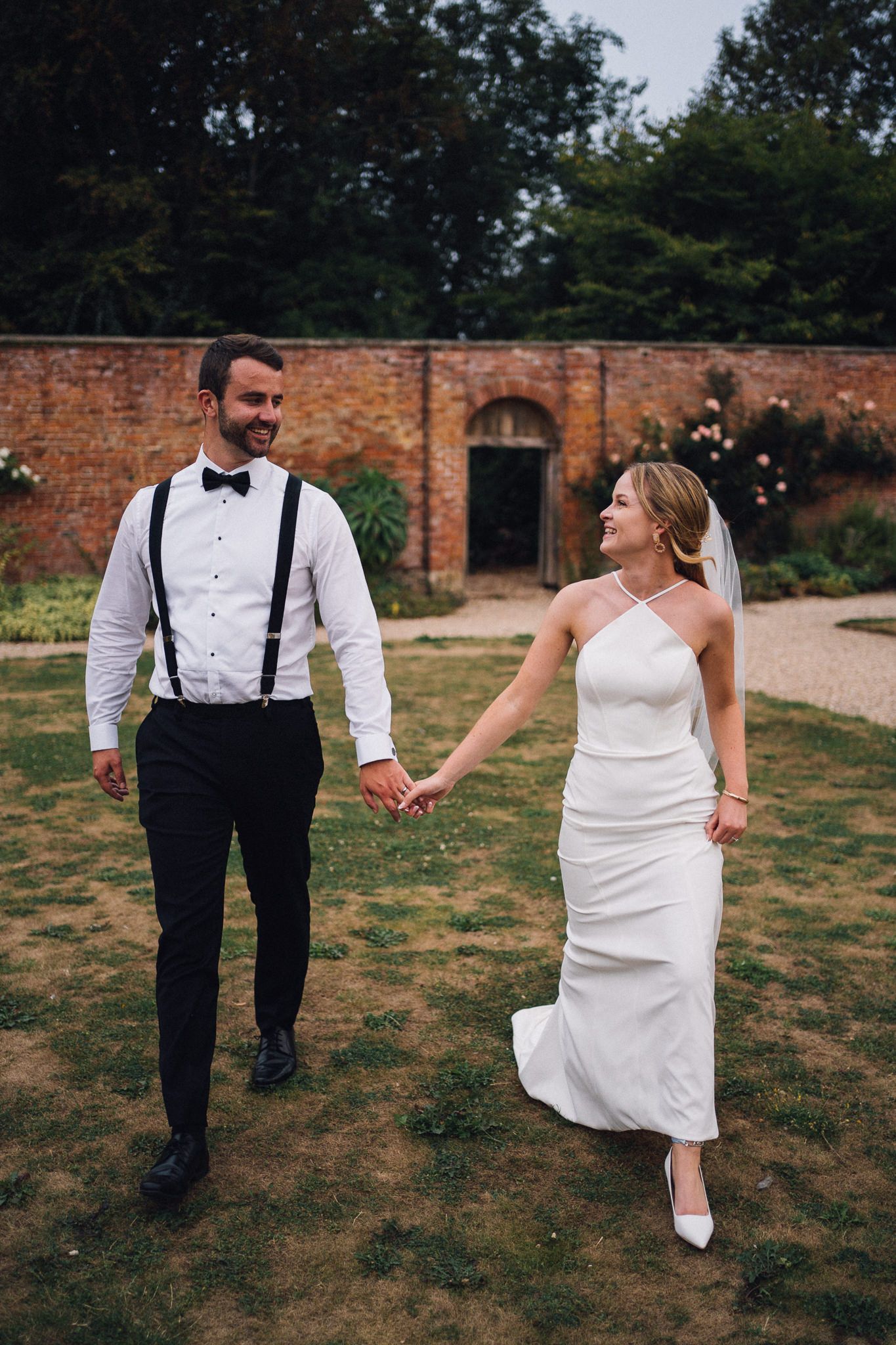A bride and groom dance playfully on a grassy lawn near a brick building.