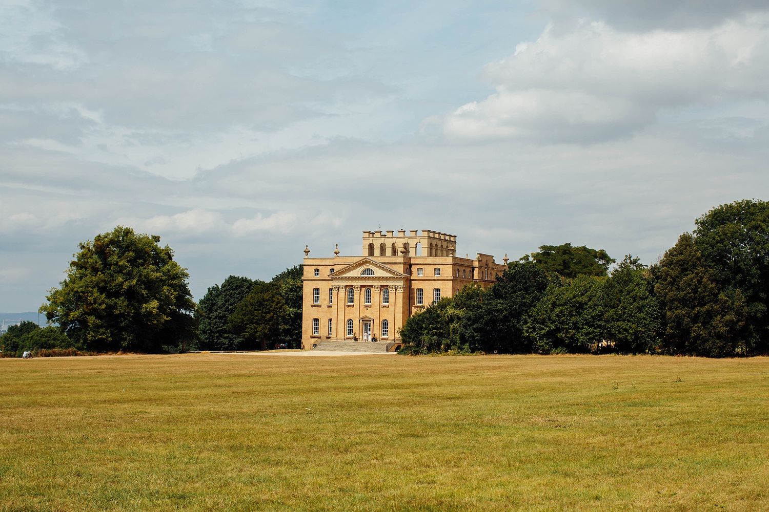 A grand historic manor house sits majestically on expansive manicured lawns under cloudy skies.
