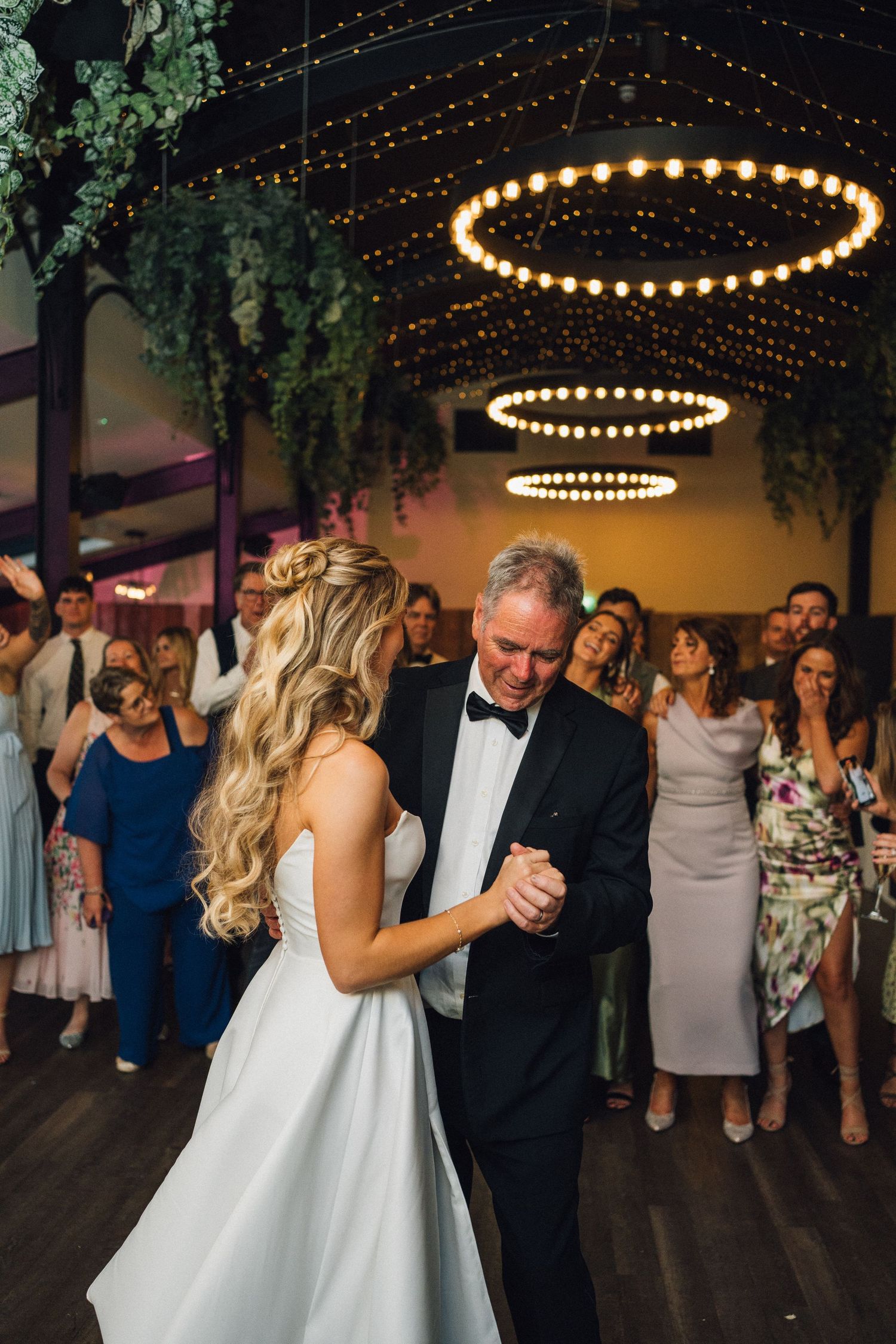Wedding reception with string lights and greenery decorations as guests dance on the floor.