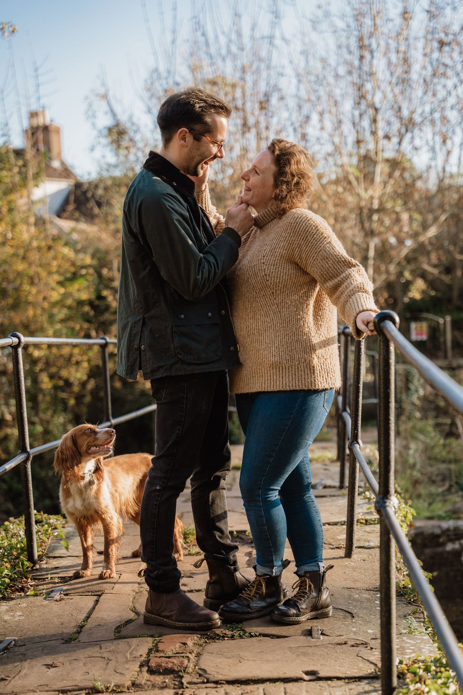 A couple shares a tender moment on a bridge walkway while their golden retriever dog stands nearby in autumn sunlight.