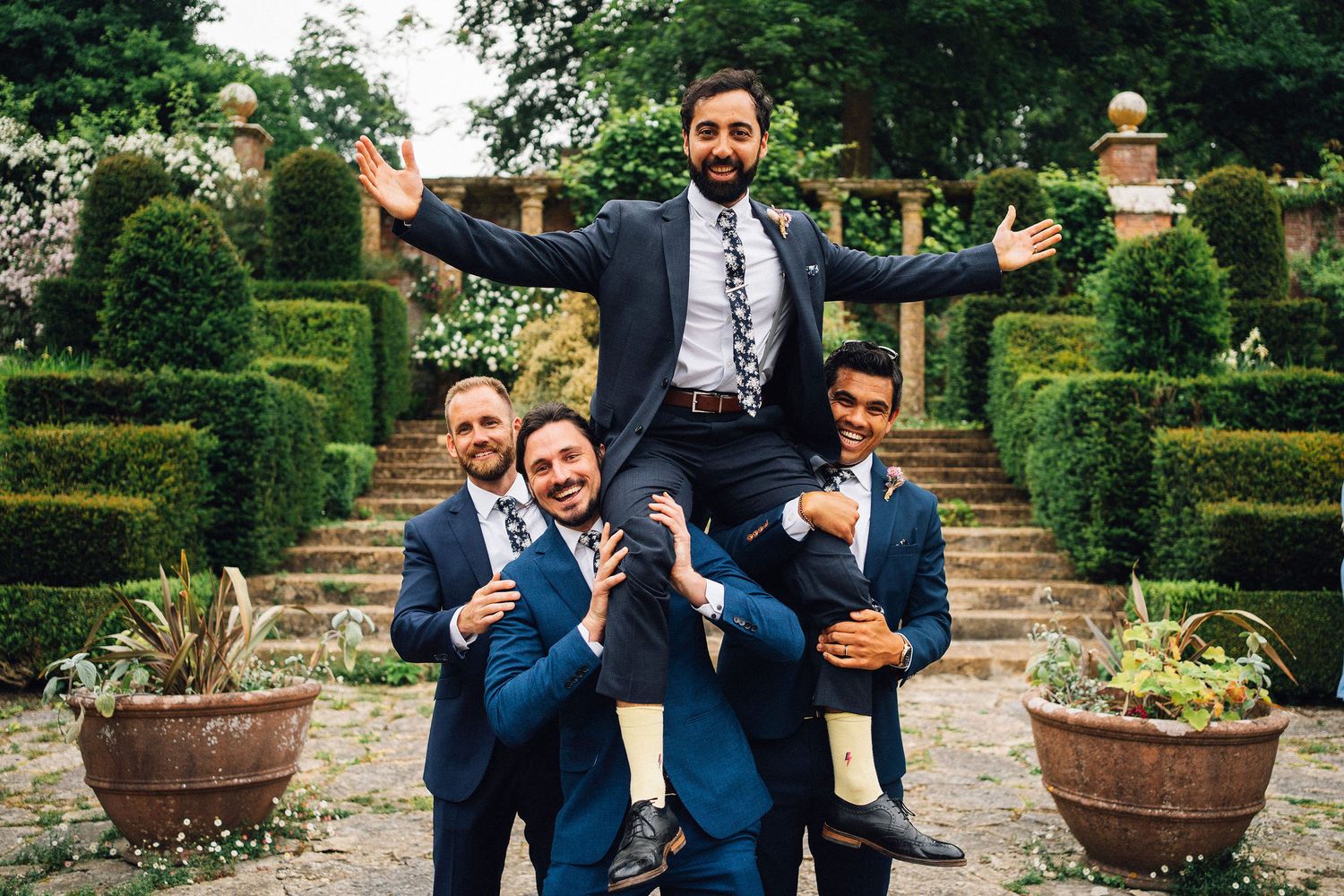 Wedding party poses playfully in a formal garden setting with manicured hedges and potted plants.