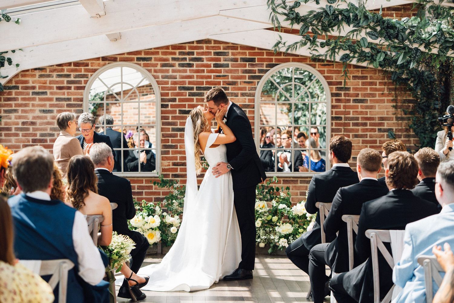 Wedding ceremony guests gather in a rustic brick courtyard adorned with greenery and white floral arrangements at Fox tail barns.
