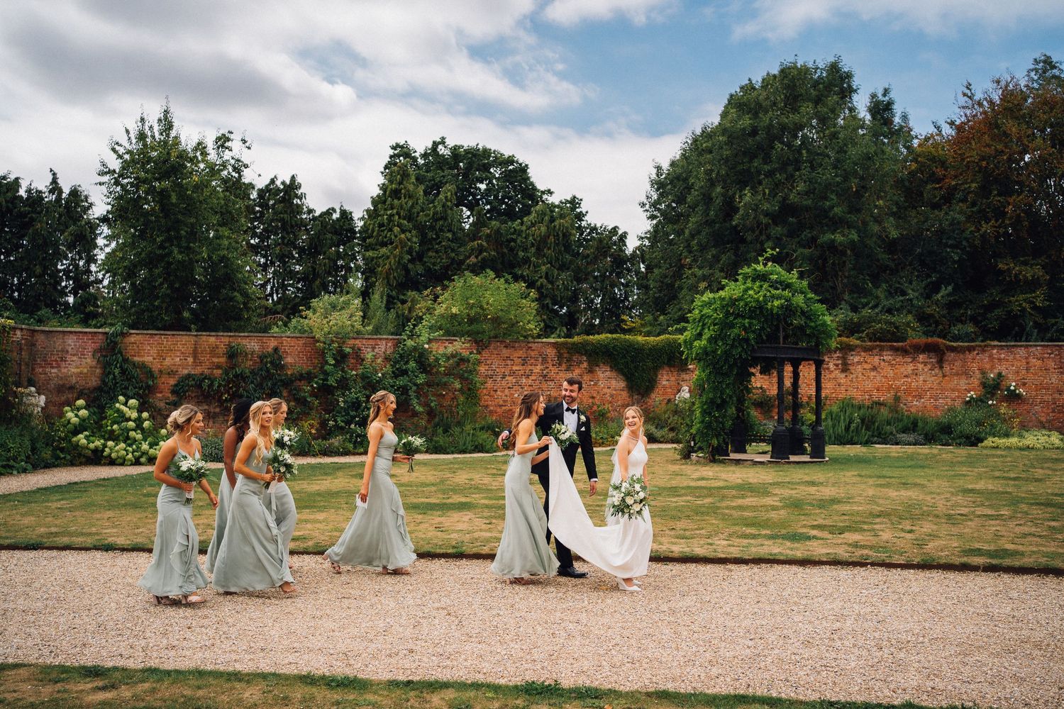 Wedding party walking through a beautiful garden with brick walls and greenery on a cloudy summer day.