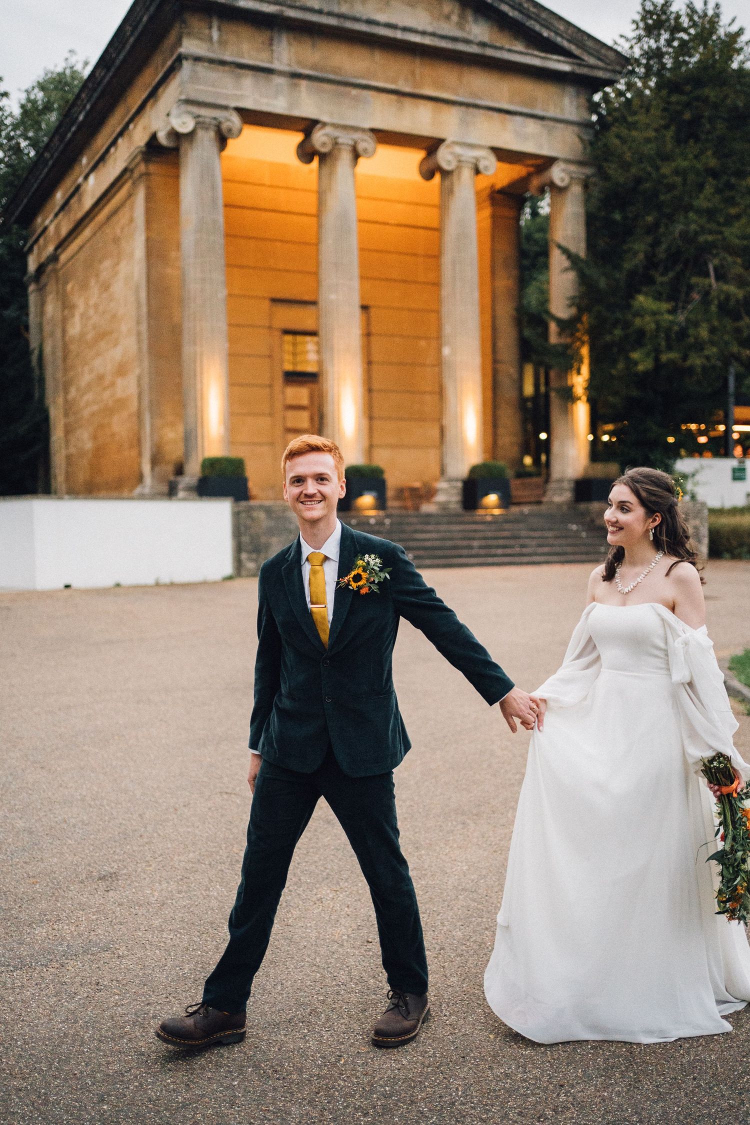 A couple in wedding attire dances and spins playfully in front of a classical stone building at sunset.