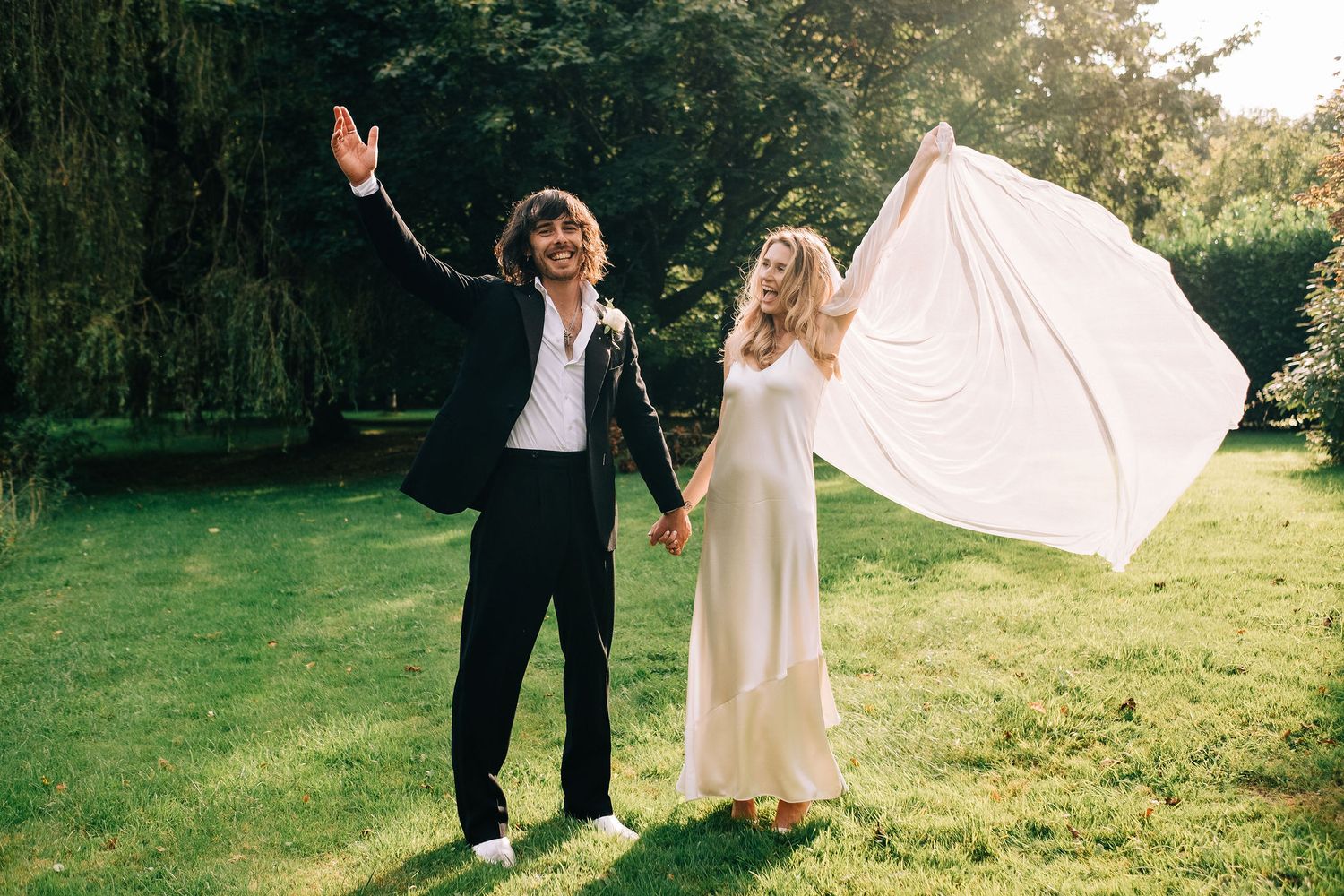 A romantic outdoor wedding photoshoot with a flowing white dress and veil blowing in the breeze near trees.