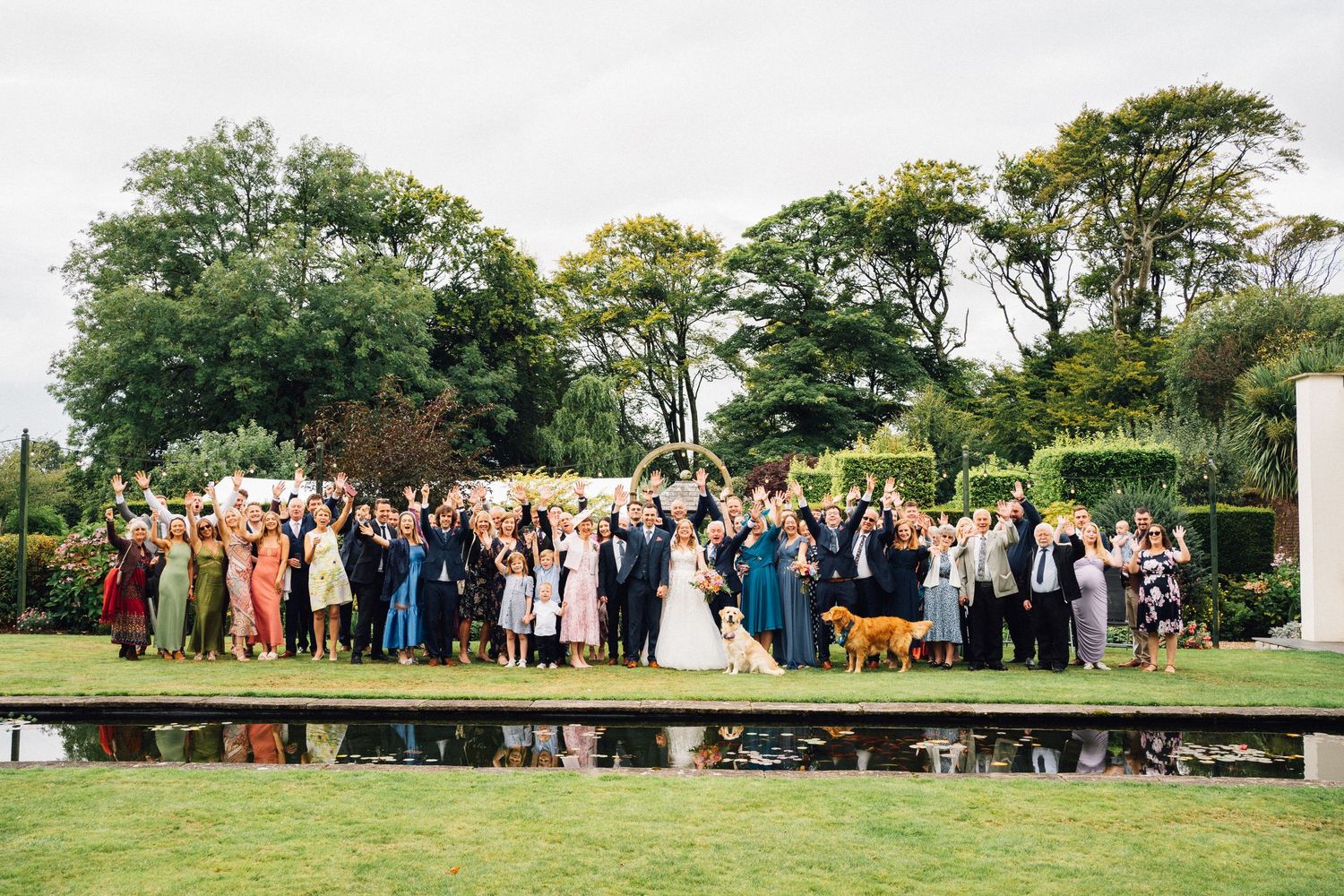 Large wedding party group photo taken outdoors on a wooden platform surrounded by trees.